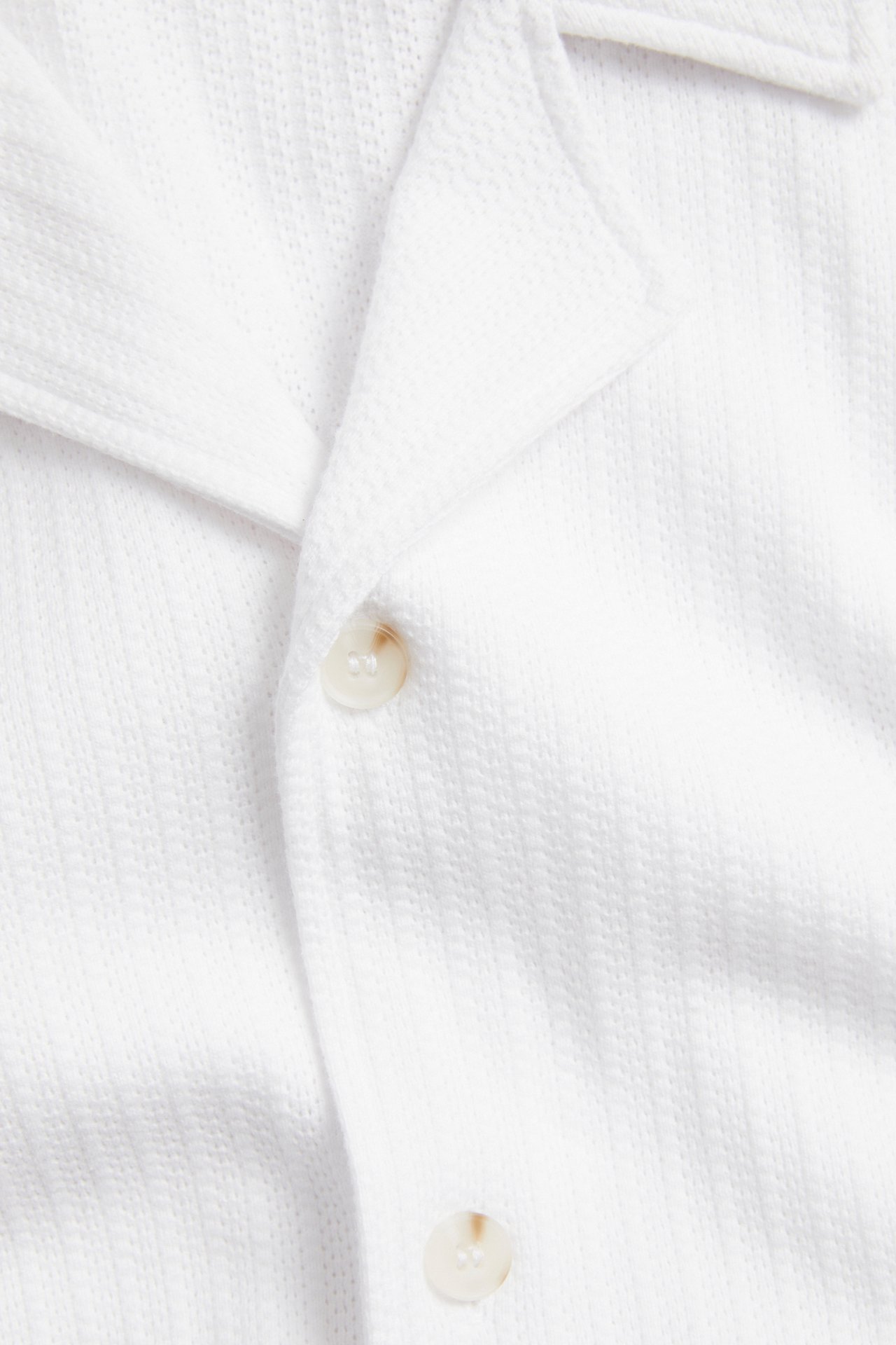 Närbild av en vit pointelle jersey pikétröja med resortkrage och två ljusa knappar.