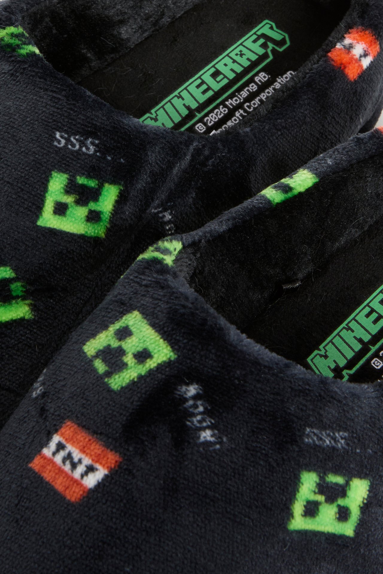 Nærbilde av svarte plysjtøfler med grønne Minecraft Creeper- og TNT-mønstre.