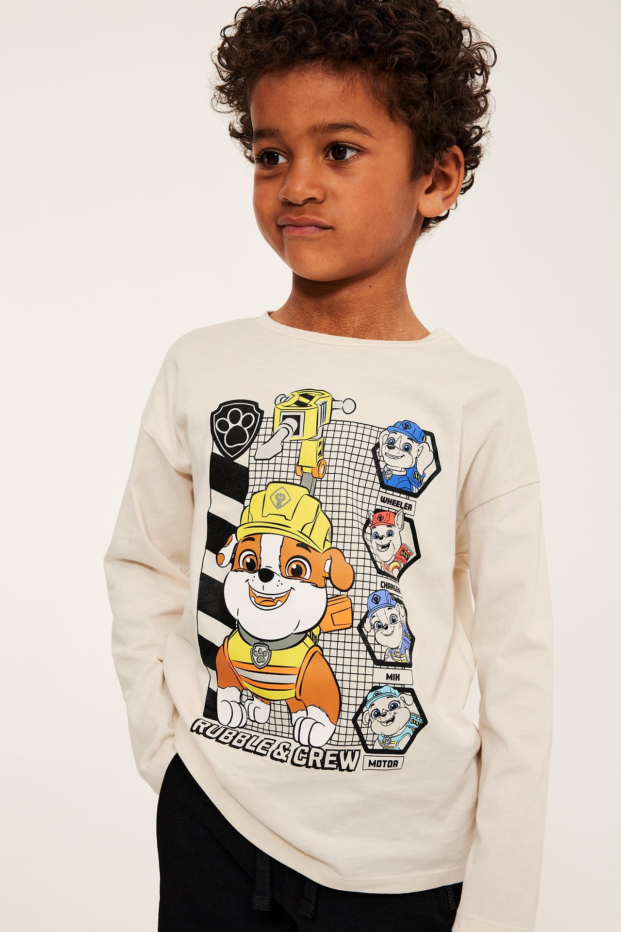 Frontbilde av et barn i en kremhvit langermet Paw Patrol-t-skjorte med trykk og svarte bukser.