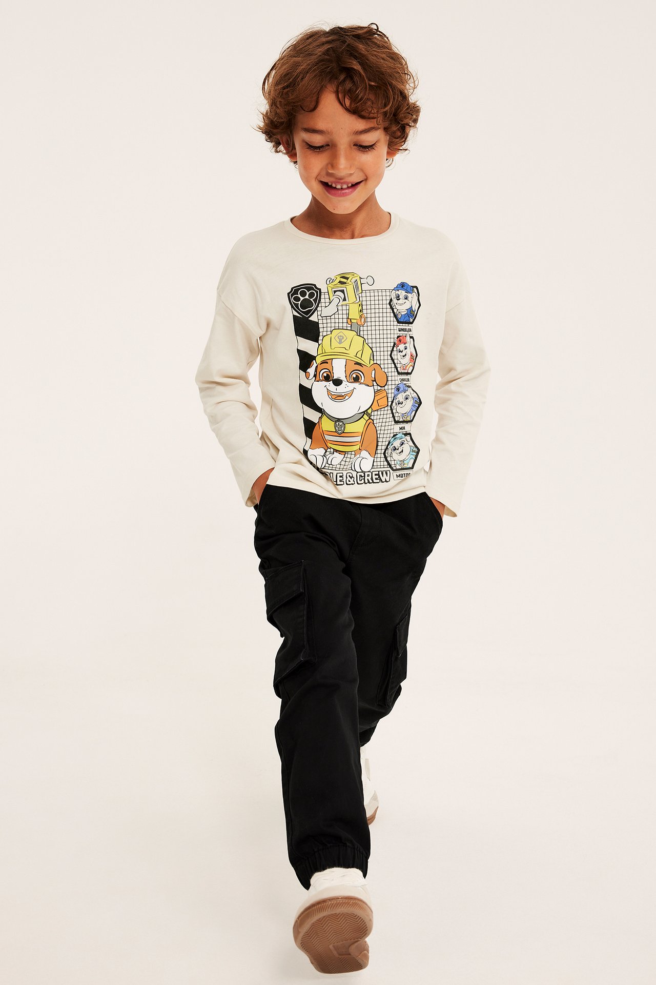 Forfra: barn i beige langermet Paw Patrol-t-skjorte med trykk, svarte cargobukser og hvite sko.