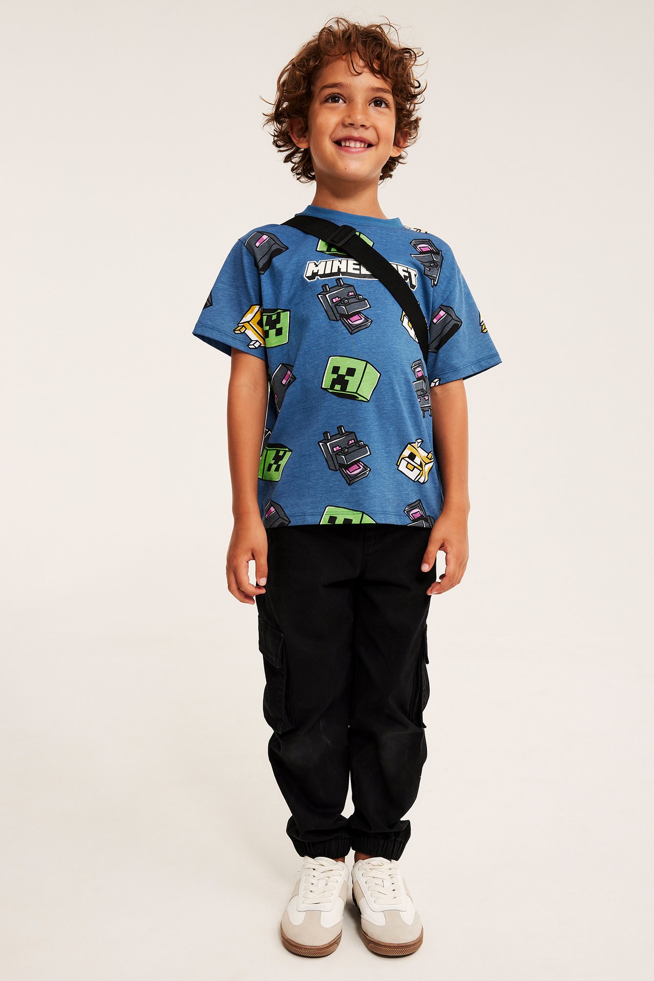 Barns blå Minecraft-mönstrad t-shirt, svart crossbody-rem, svarta cargobyxor, vita sneakers.