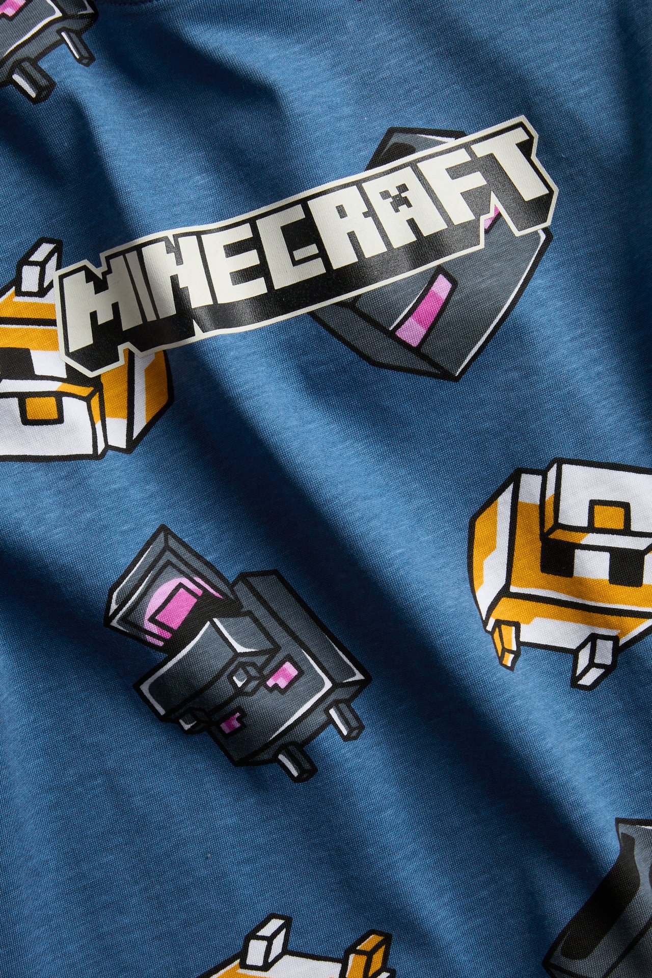 Zbliżenie na niebieską koszulkę z białym logo Minecraft i różnymi pikselowymi grafikami z gry.