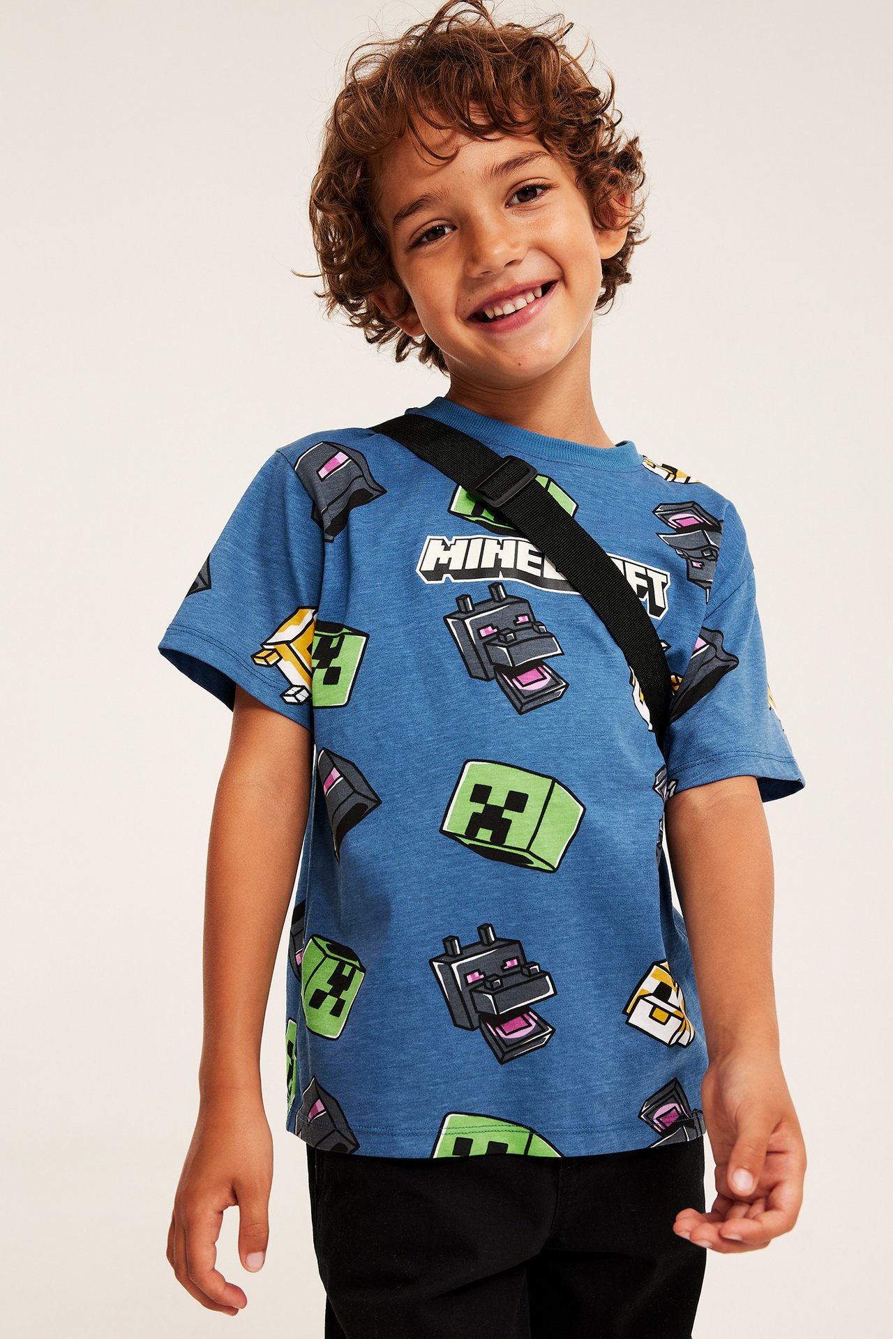 Frontvy av ett barn i en blå kortärmad t-shirt med Minecraft-tryck, svart axelrem och svarta byxor.