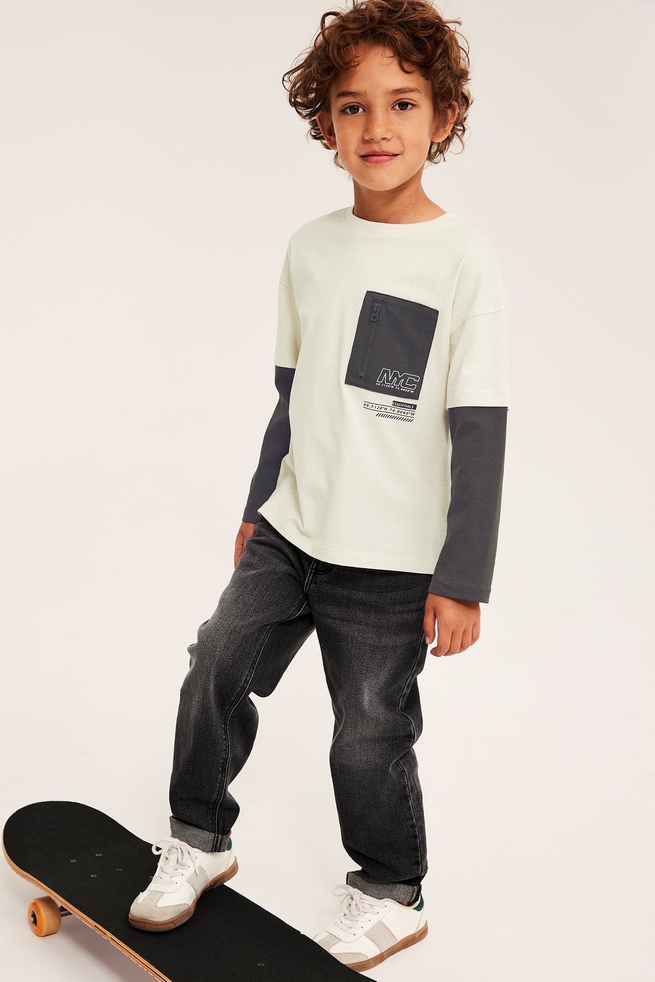 Pojks långärmad tröja i benvitt och mörkgrått med bröstficka, mörkgrå jeans, vita sneakers.