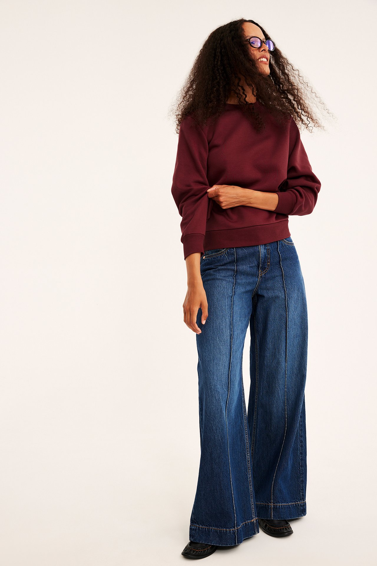 Frontbilde av kvinnelig modell i blå vide jeans, en vinrød genser og mørke loafers.