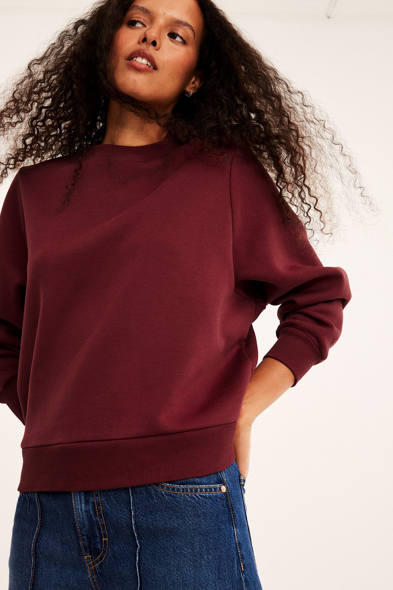Front av kvinne i vinrød crewneck genser med ribbekant og blå jeans.