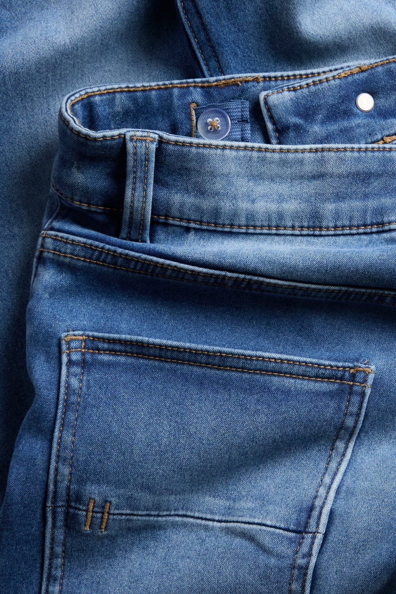 Zbliżenie na niebieskie jeansy z denimu z pomarańczowymi szwami, przezroczystym guzikiem i tylną kieszenią.