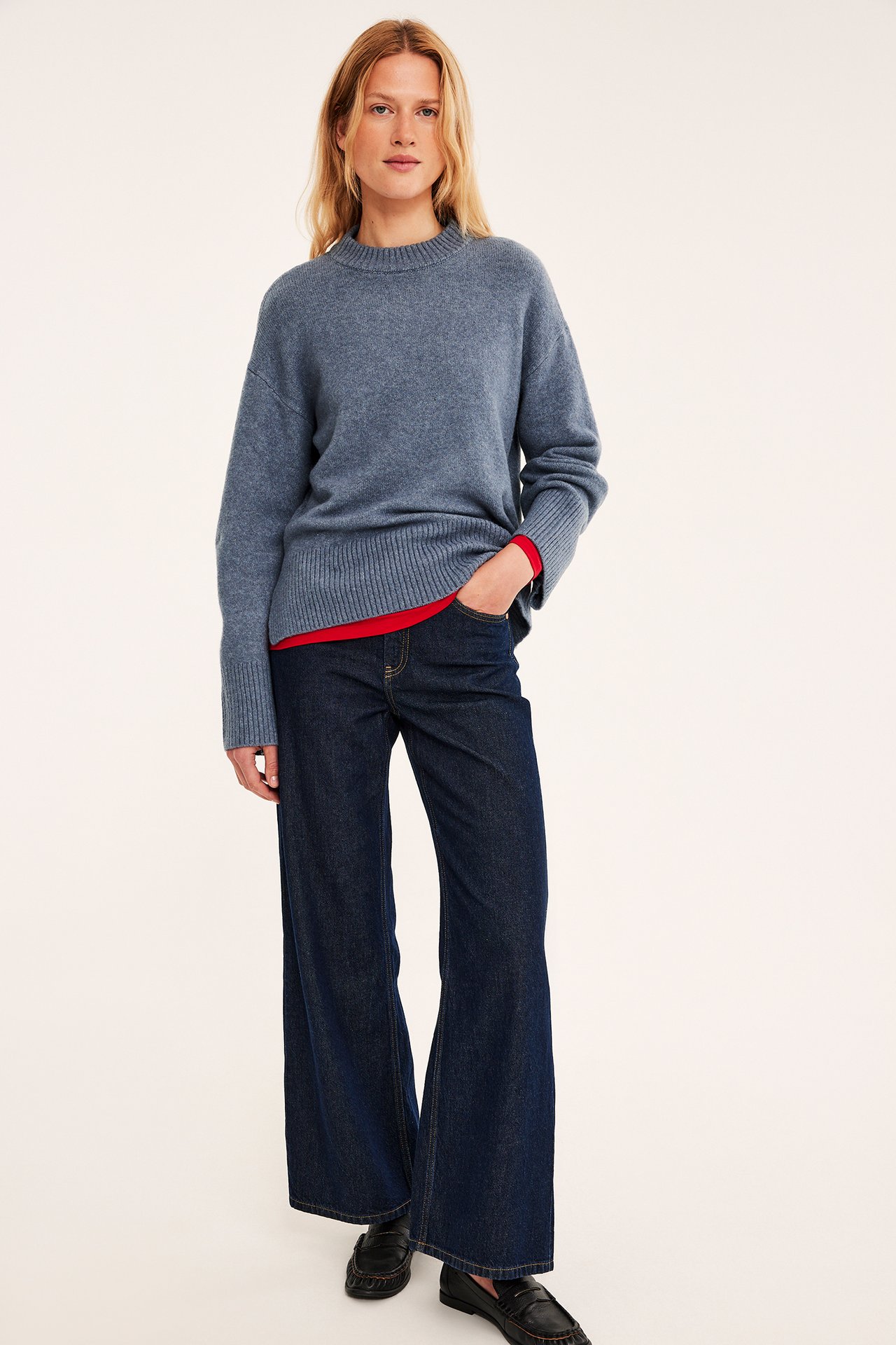 Kvinne forfra i blå genser over rød skjorte, mørkeblå vide jeans, svarte loafers.
