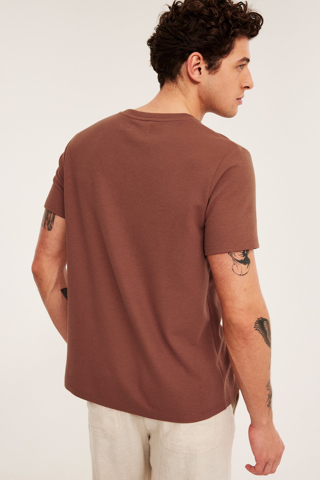 Widok od tyłu: mężczyzna w brązowej teksturowanej koszulce regular fit i jasnobeżowych spodniach.