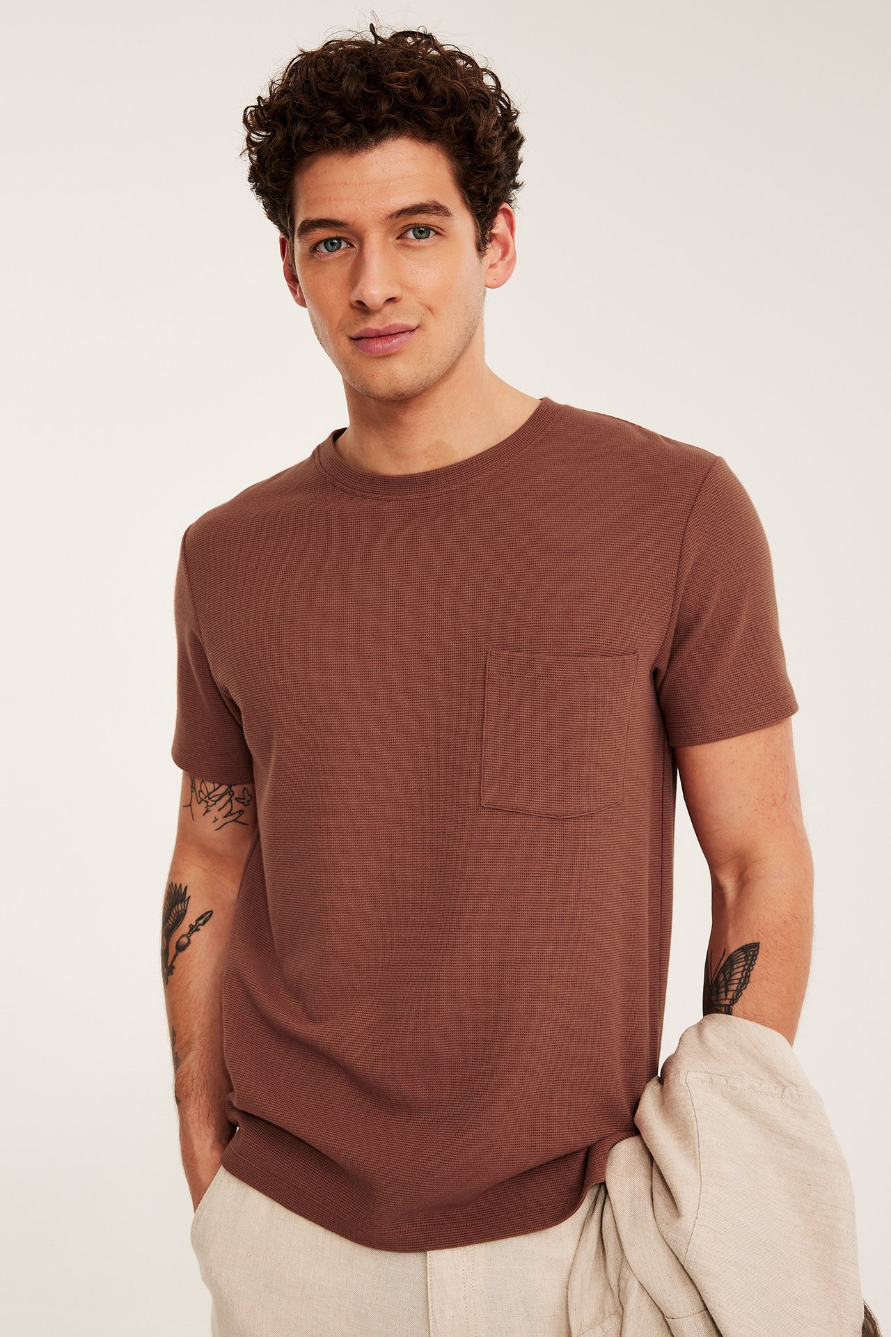 Mężczyzna w brązowym, teksturowanym T-shircie z kieszenią, jasnobeżowych spodniach i kurtce.