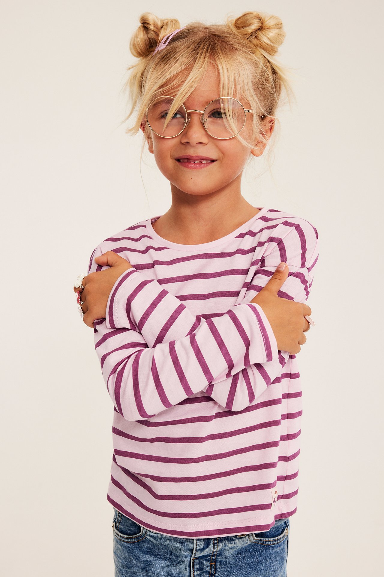 Rosa og lilla stripet topp på et barn, med blå jeans, runde briller og ringer.