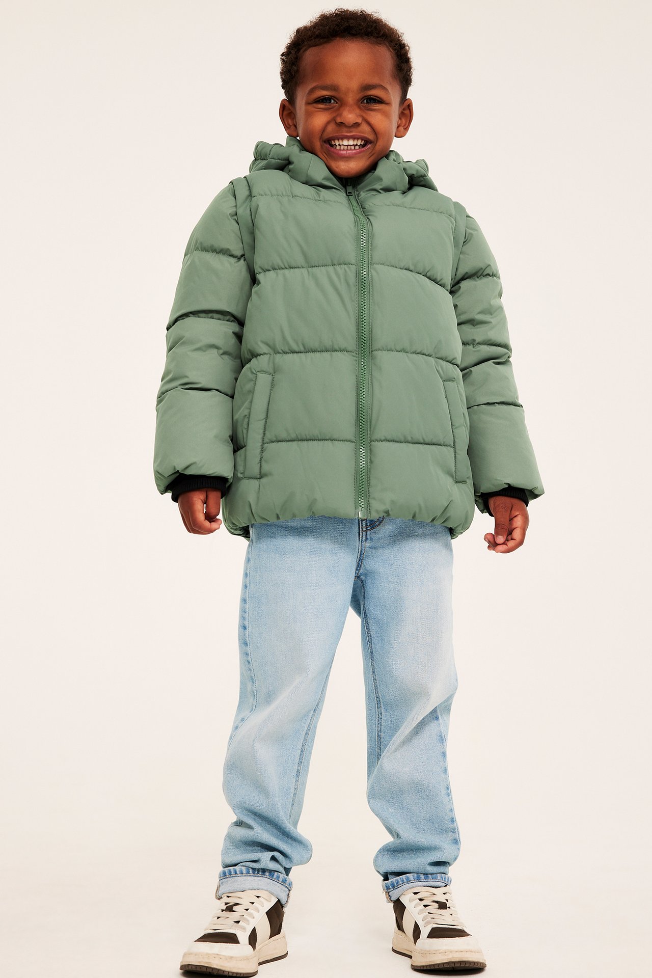 Framifrån: ett barn i grön vadderad jacka med huva, ljusblå jeans och svartvita sneakers.