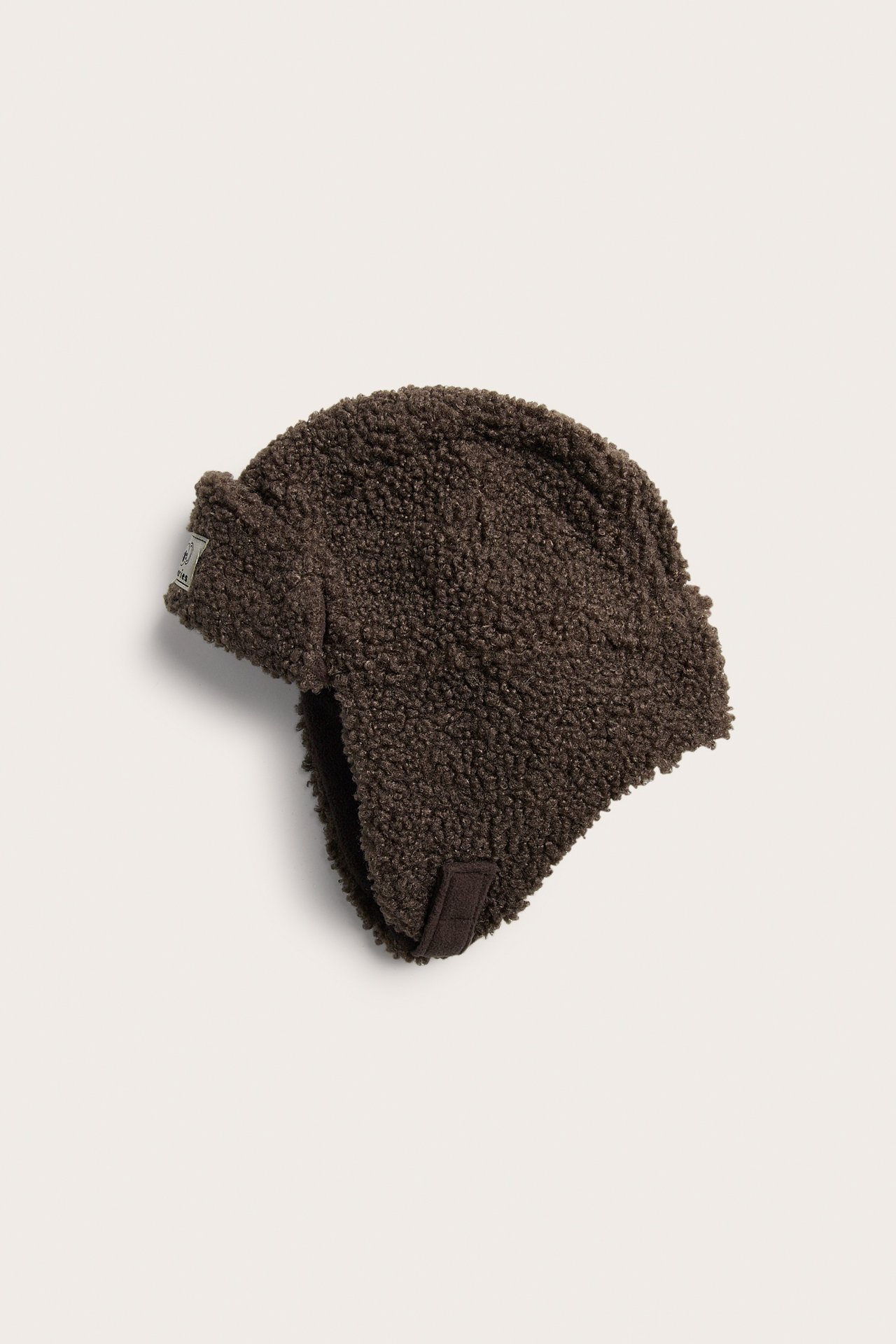 Vinklad toppvy av en mörkbrun teddyfleece-mössa med öronlappar och en rem.