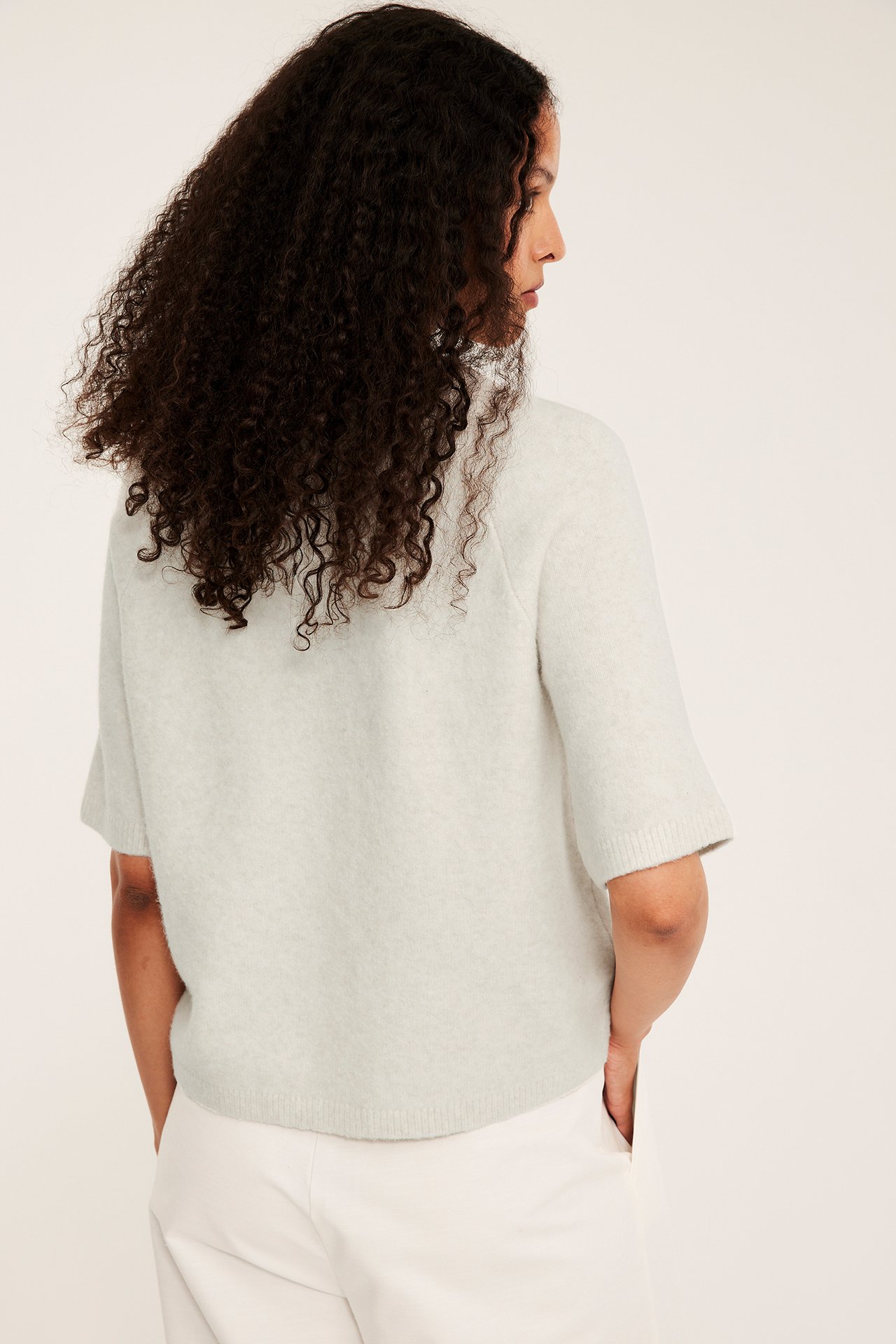 Bakre sidovy av en kvinna i en oversize beige melerad stickad kortärmad t-shirt och vita byxor.