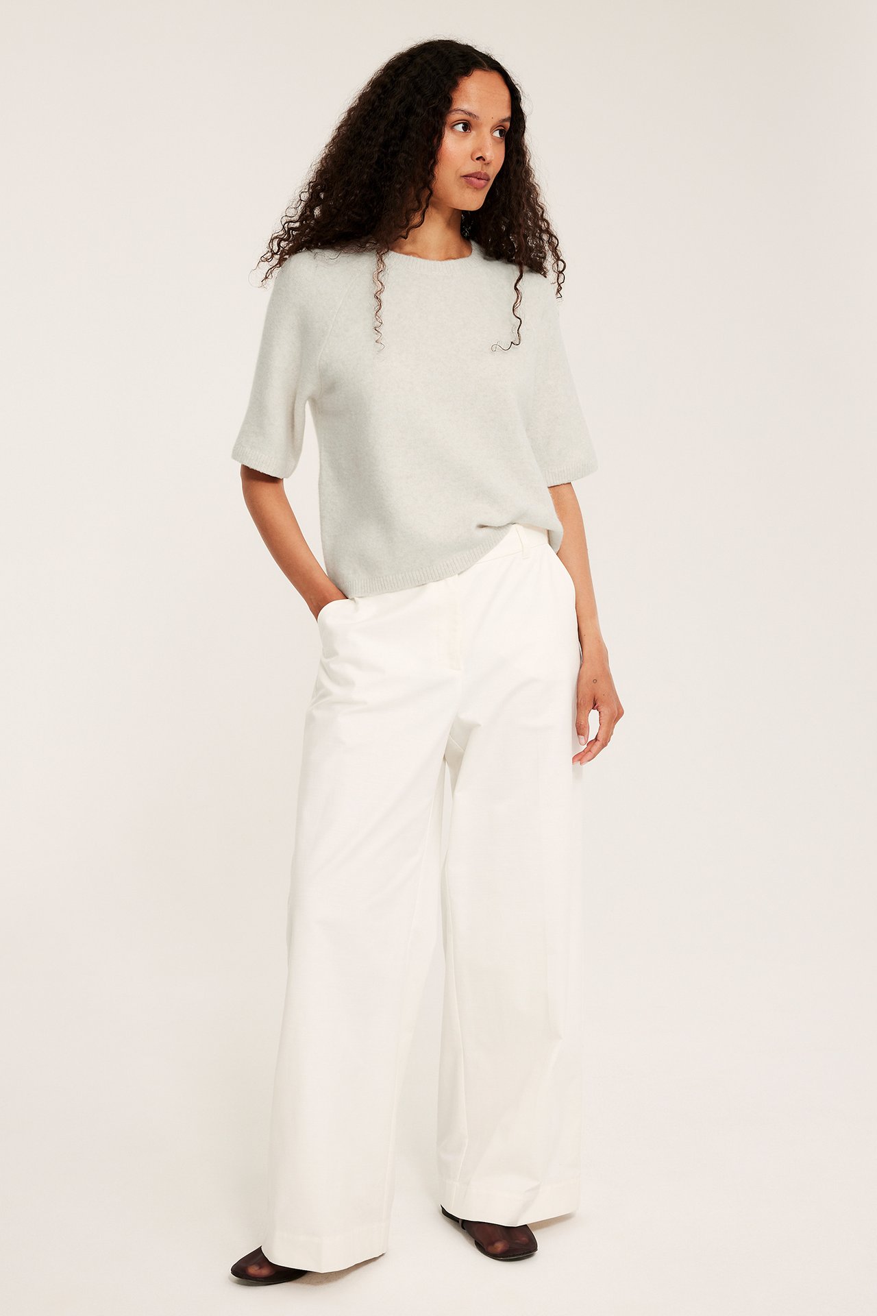 Frontvy av kvinna i oversized melerad beige stickad t-shirt och vita vida byxor.