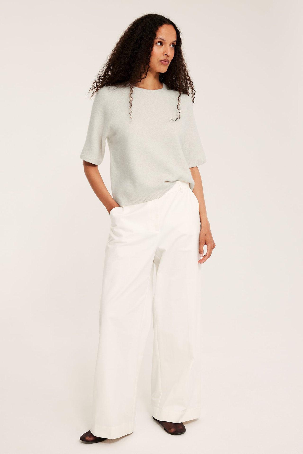 Frontvy av kvinna i oversized melerad beige stickad t-shirt och vita vida byxor.