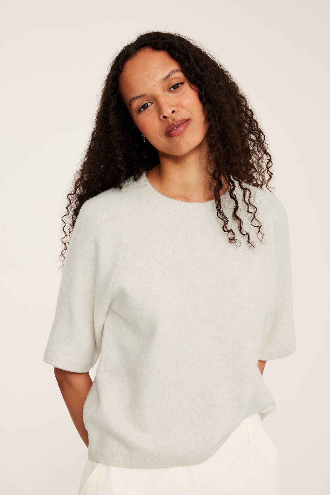 Framsida: kvinna i oversized, kortärmad, melerad beige stickad t-shirt och vita byxor.