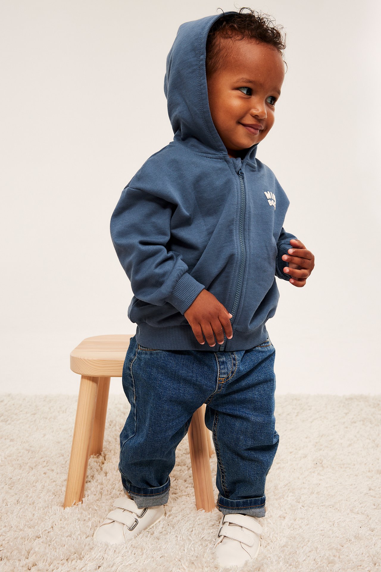 Framifrån, baby i blå huvtröja med dragkedja och tryck, blå jeans och vita sneakers.
