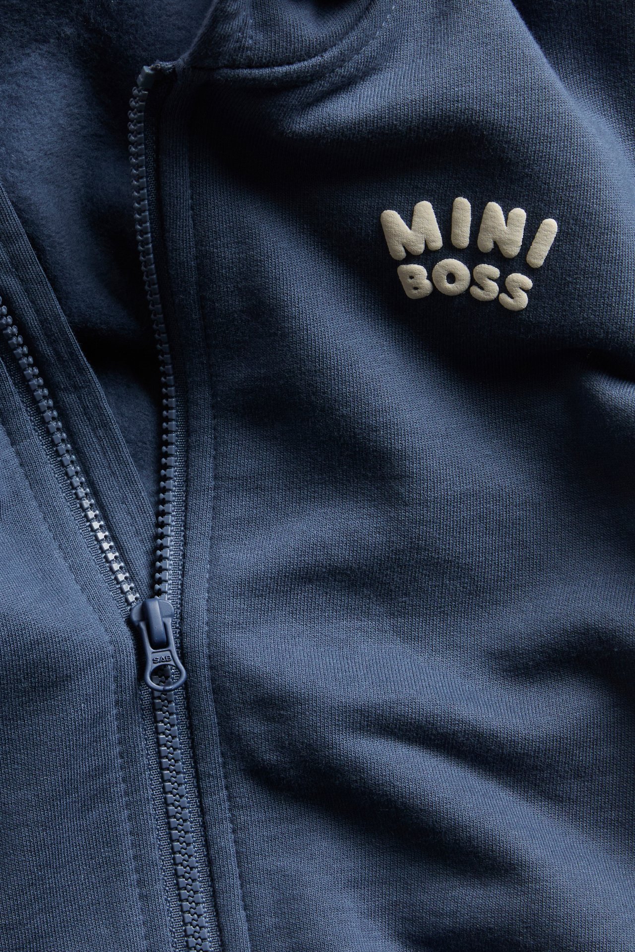 Närbild på en blå barnhoodie med texten 'MINI BOSS' och dragkedja framtill.