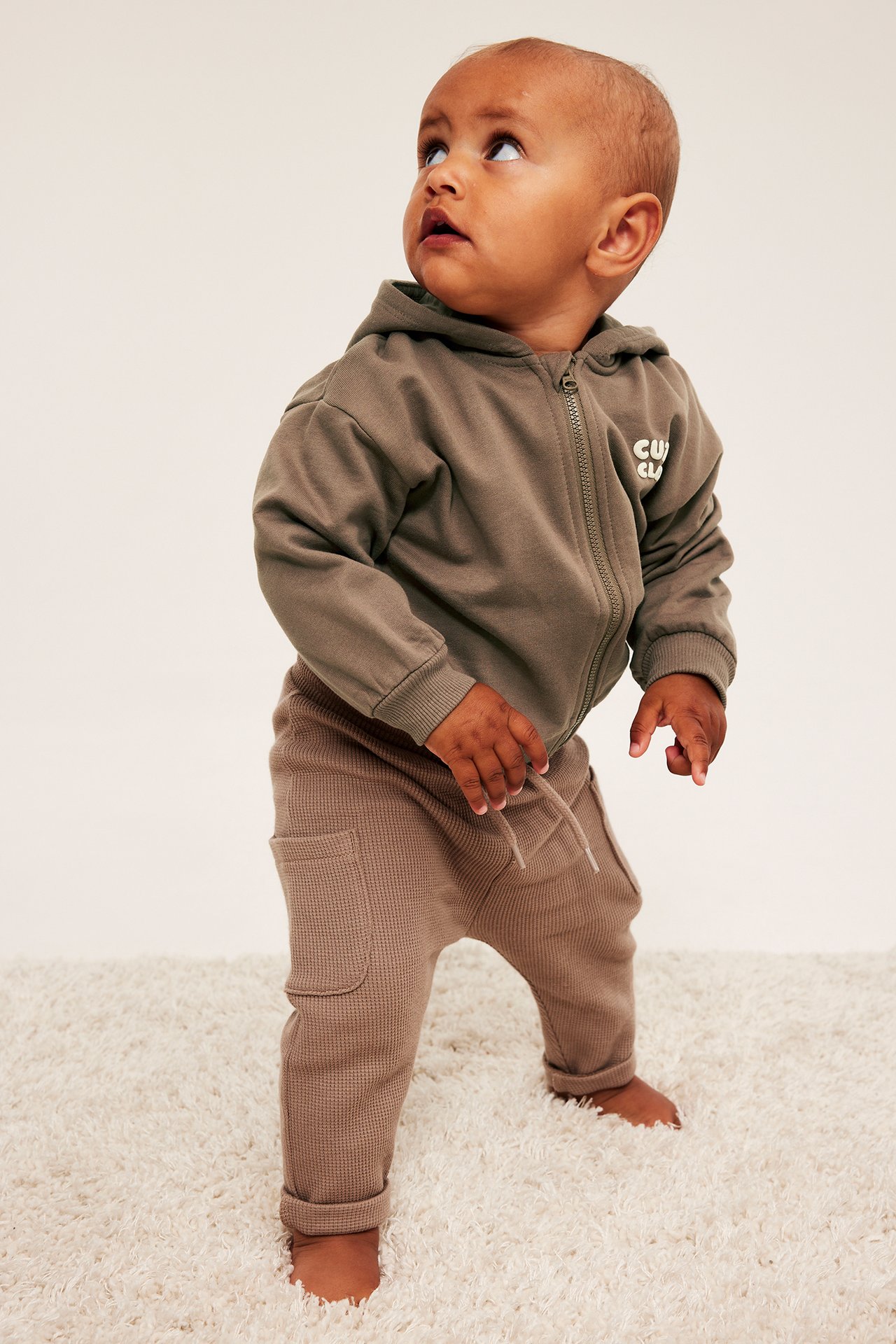 Framifrån på baby i khaki dragkedjehoodie med 'CUDD CLUB'-tryck och bruna våfflade joggare.