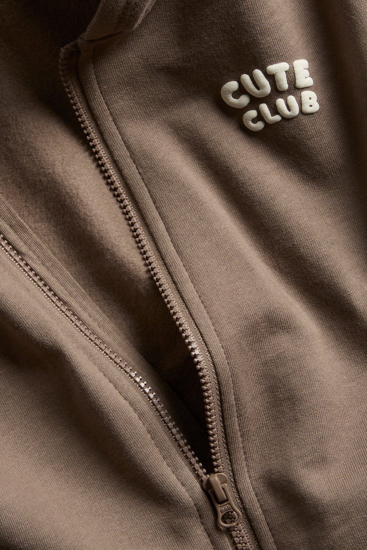 Närbild på en brun huvtröja med dragkedja, "Cute Club"-märke och fickor med dragkedja.
