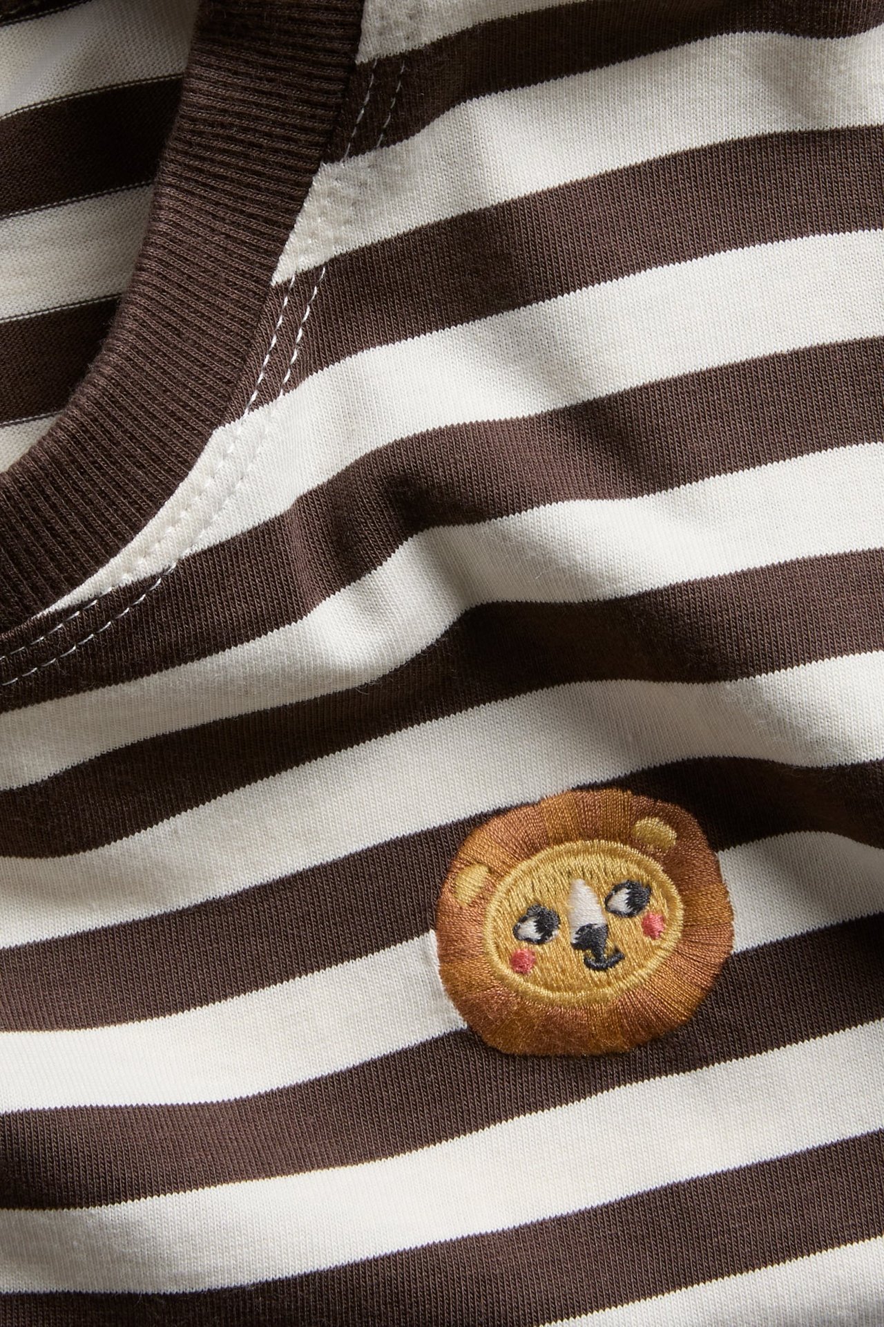 Nærbilde av en brun og hvit stripete topp med ribbet hals og en brodert løvepatch.