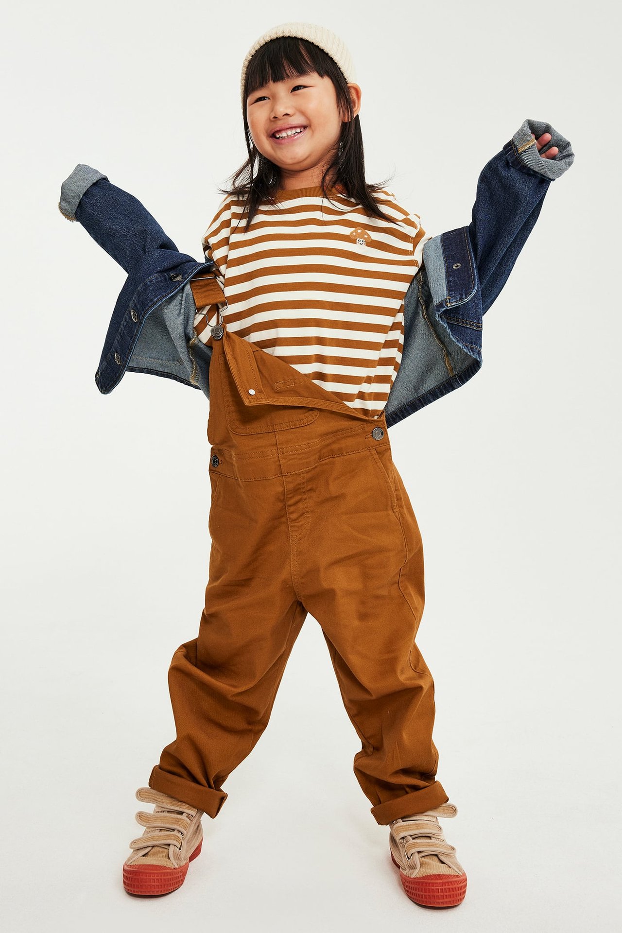 Front, barn i brune snekkerbukser, stripete t-skjorte, denimjakke, kremfarget lue og joggesko.