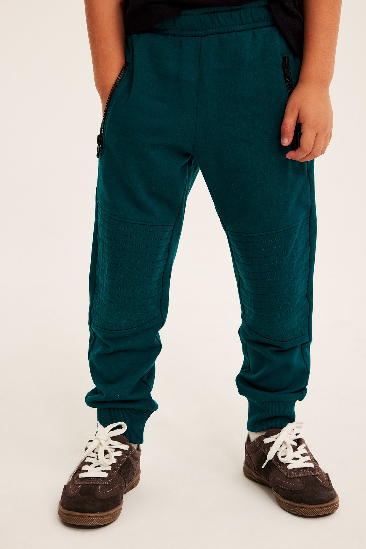 Framifrån: Barnens teal-joggingbyxor med dragkedjefickor, ribbade knän och bruna sneakers.