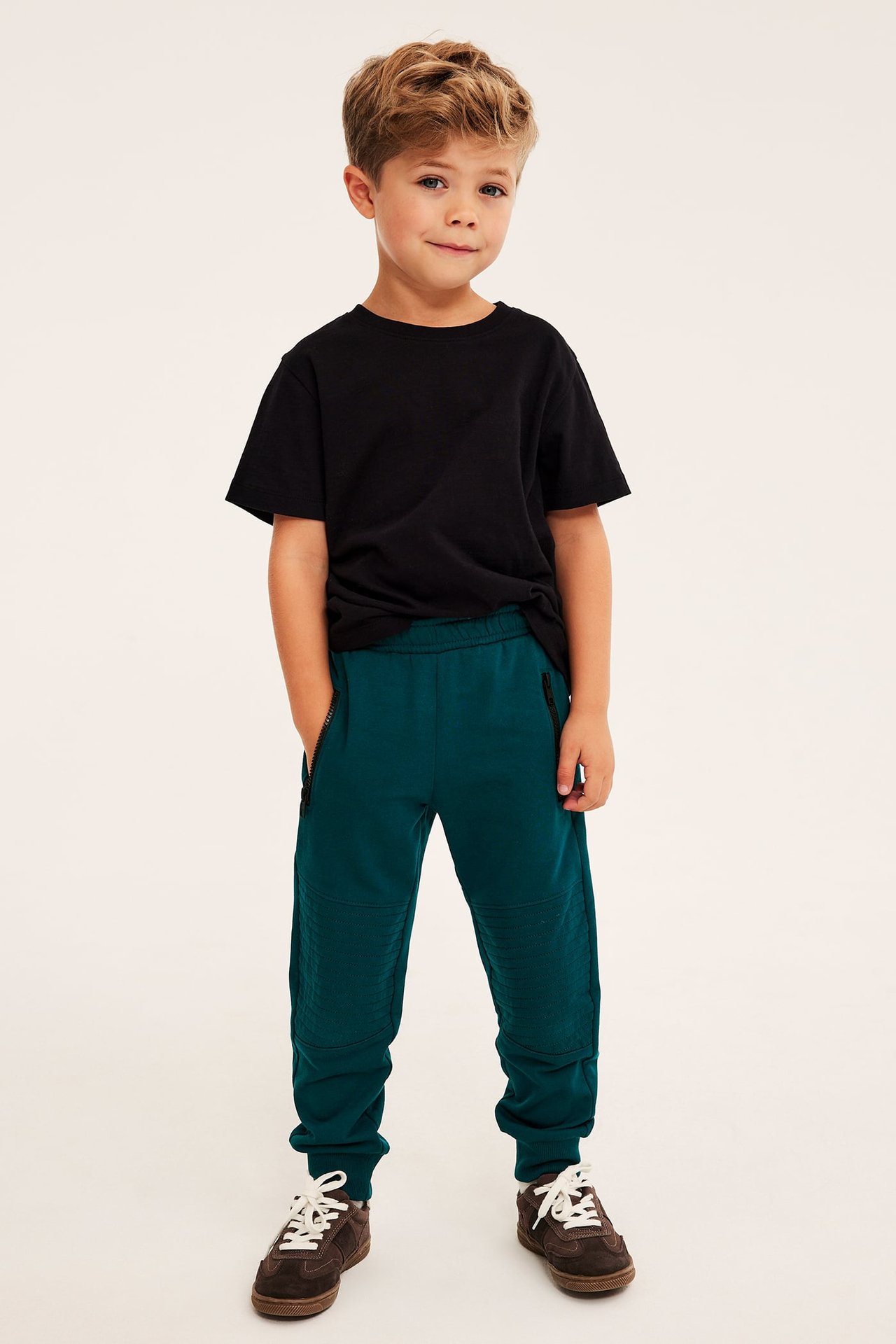 Framifrån: pojke i mörkgröna ribbade sweatpants, svart t-shirt och bruna sneakers.