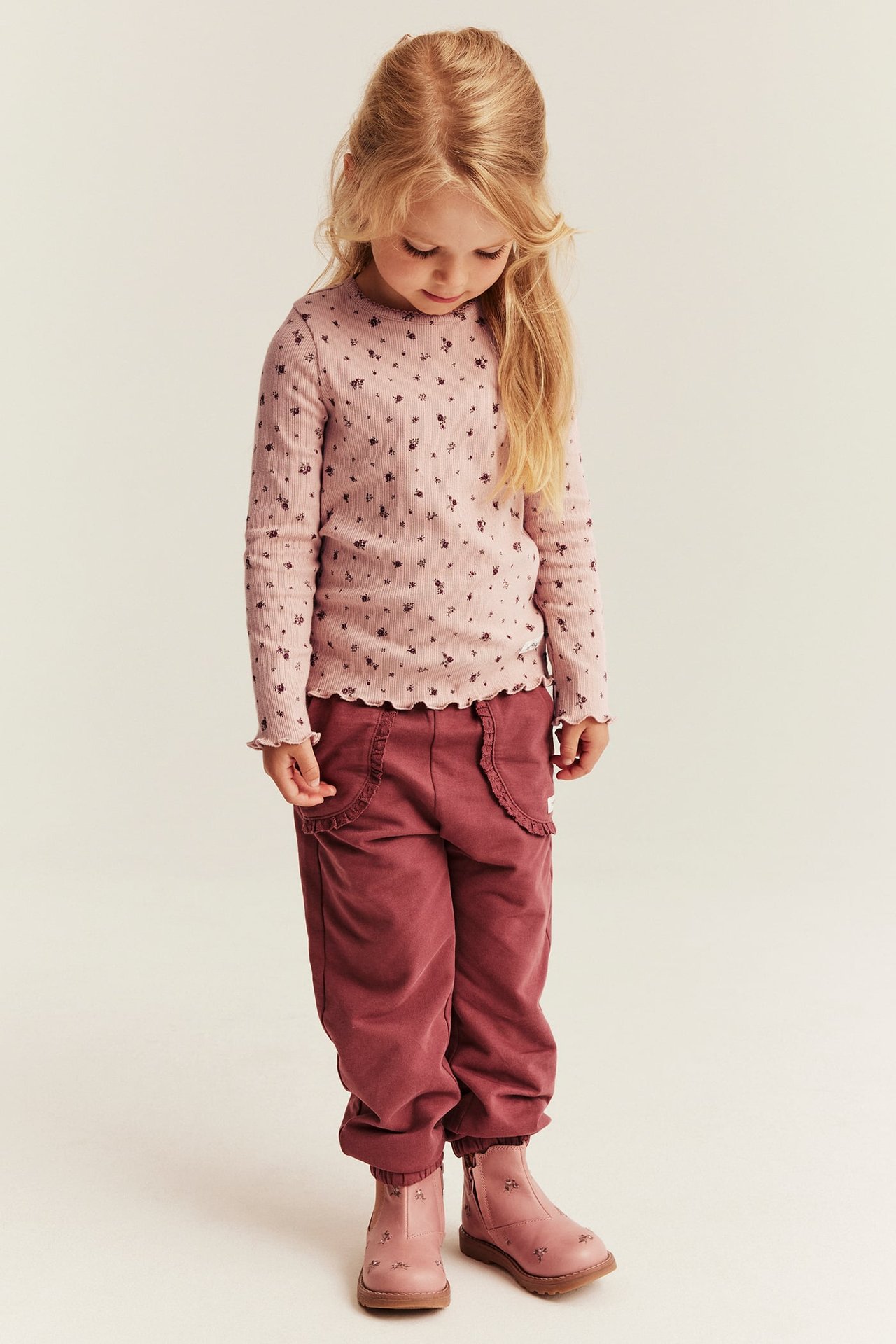 Front view, kid i rosa blommiga ribbad långärmad topp, vinröda mjukisbyxor och rosa broderade stövlar.