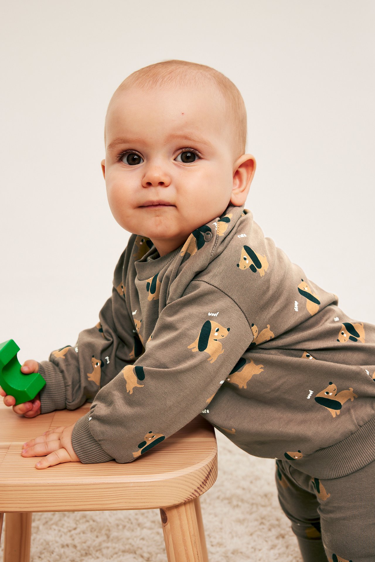 Baby i långärmad khakifärgad topp med taxmönster och matchande byxor, håller grön kloss.