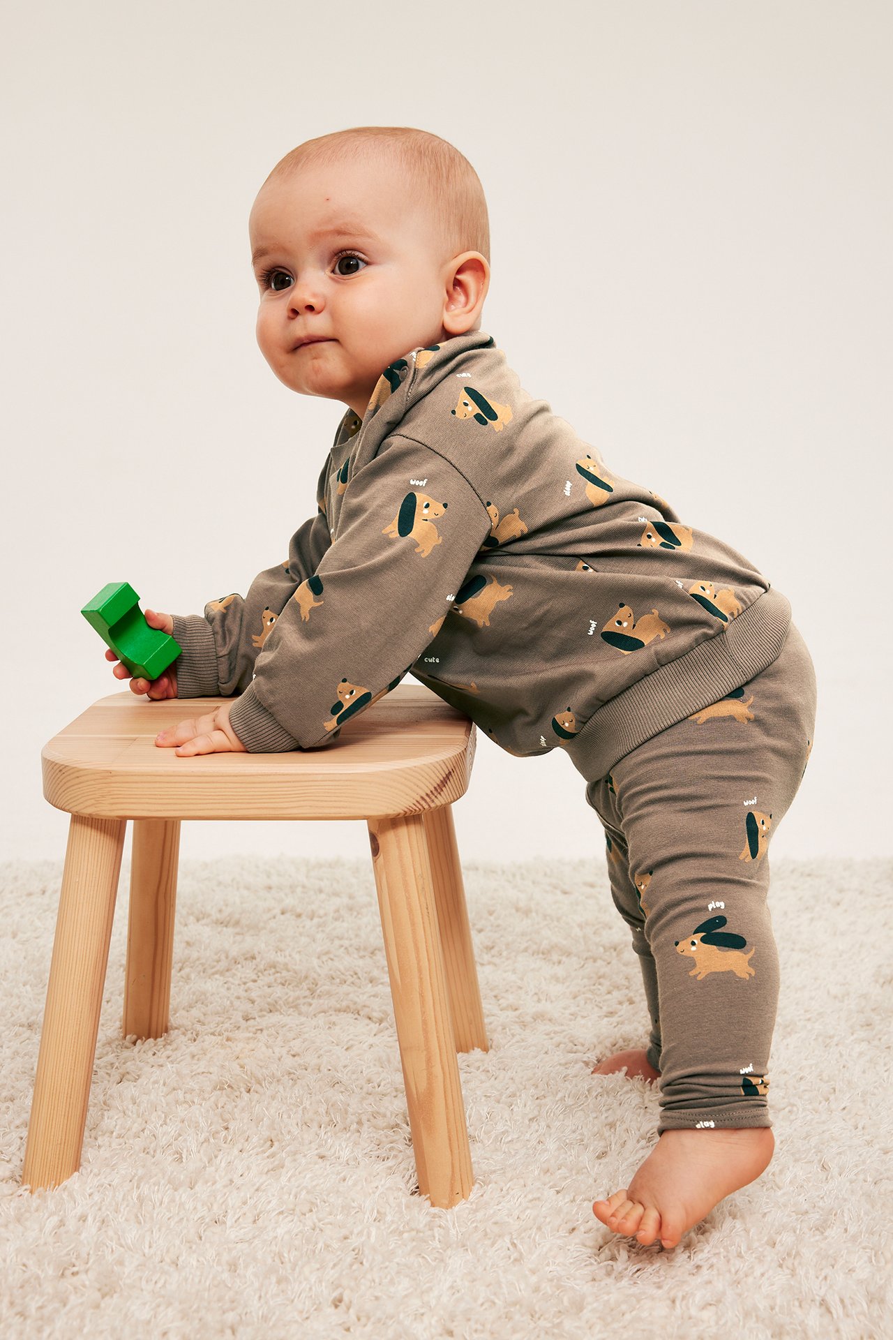 Sidovy av baby i brunmelerad tröja och matchande leggings med taxprint, håller grön kloss.