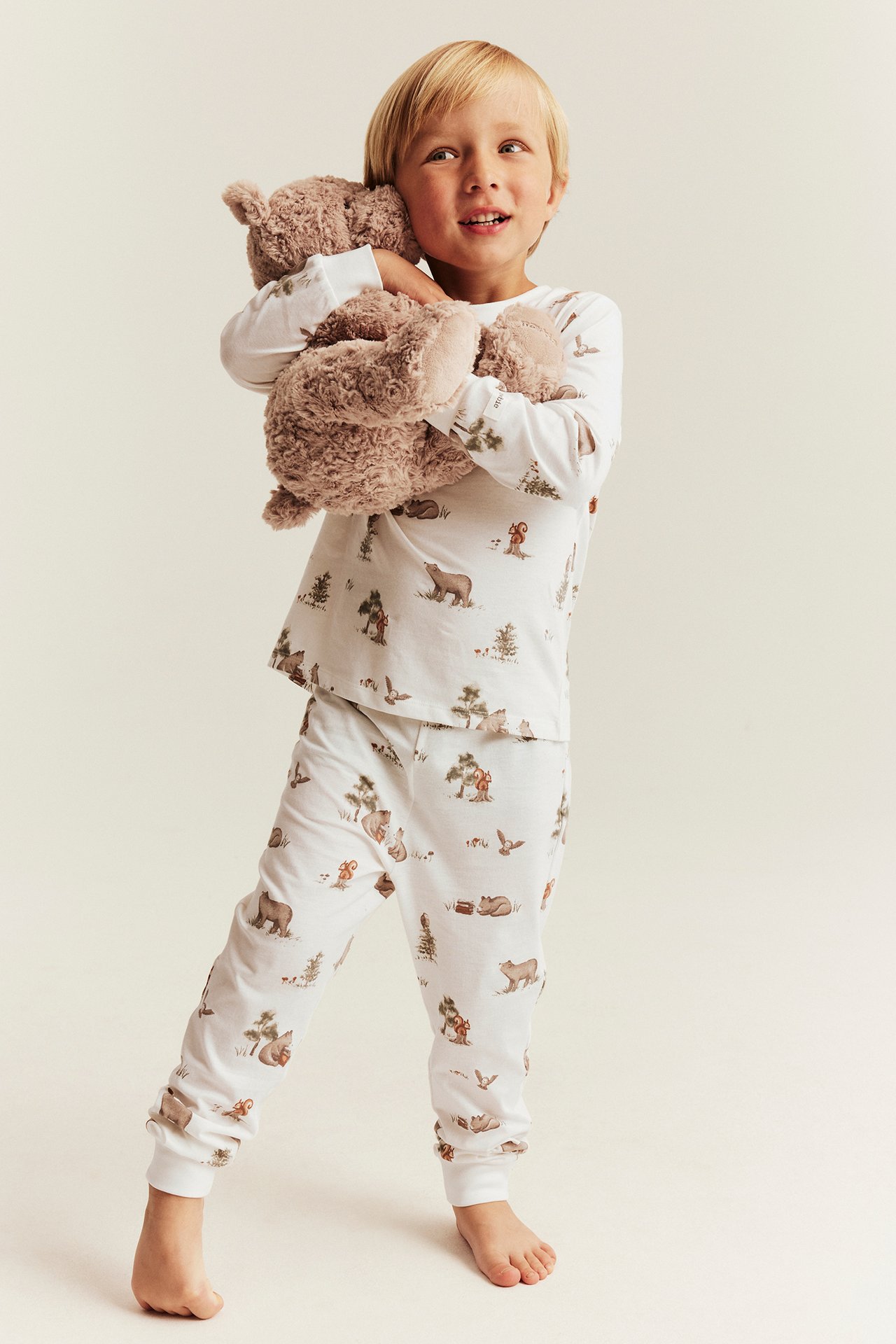 Edestä: Lapsi valkoisessa metsäneläinaiheisessa pyjamassa, pitää ruskeaa nallea.