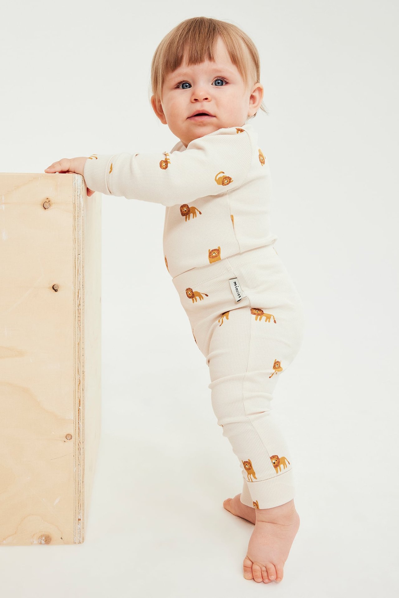 Sidebilde av en baby i et beige ribbet sett med løveprint, holder en trekasse.