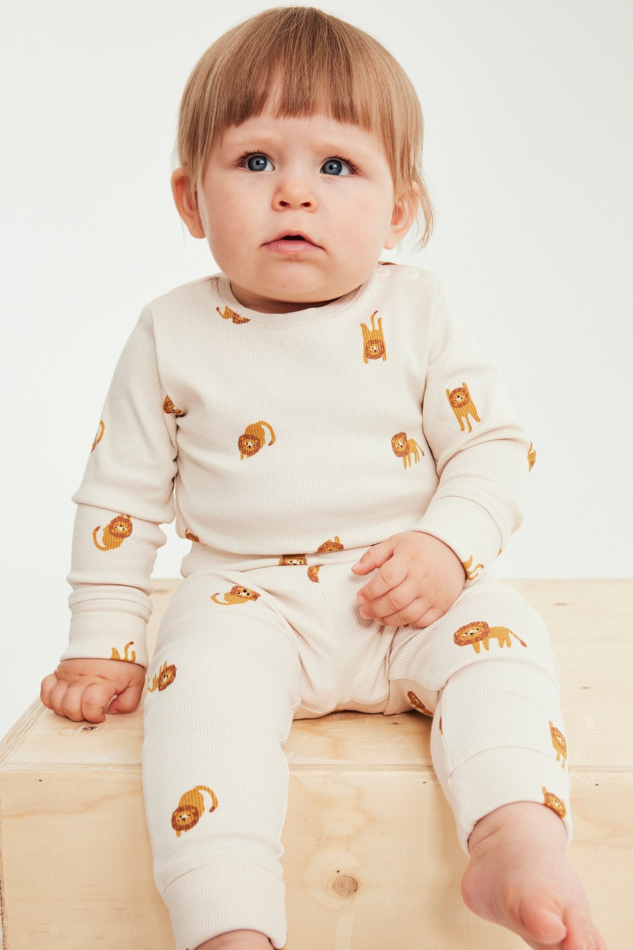 Frontvisning av baby i lys beige ribbestrikket langermet topp og bukse med løvetrykk.