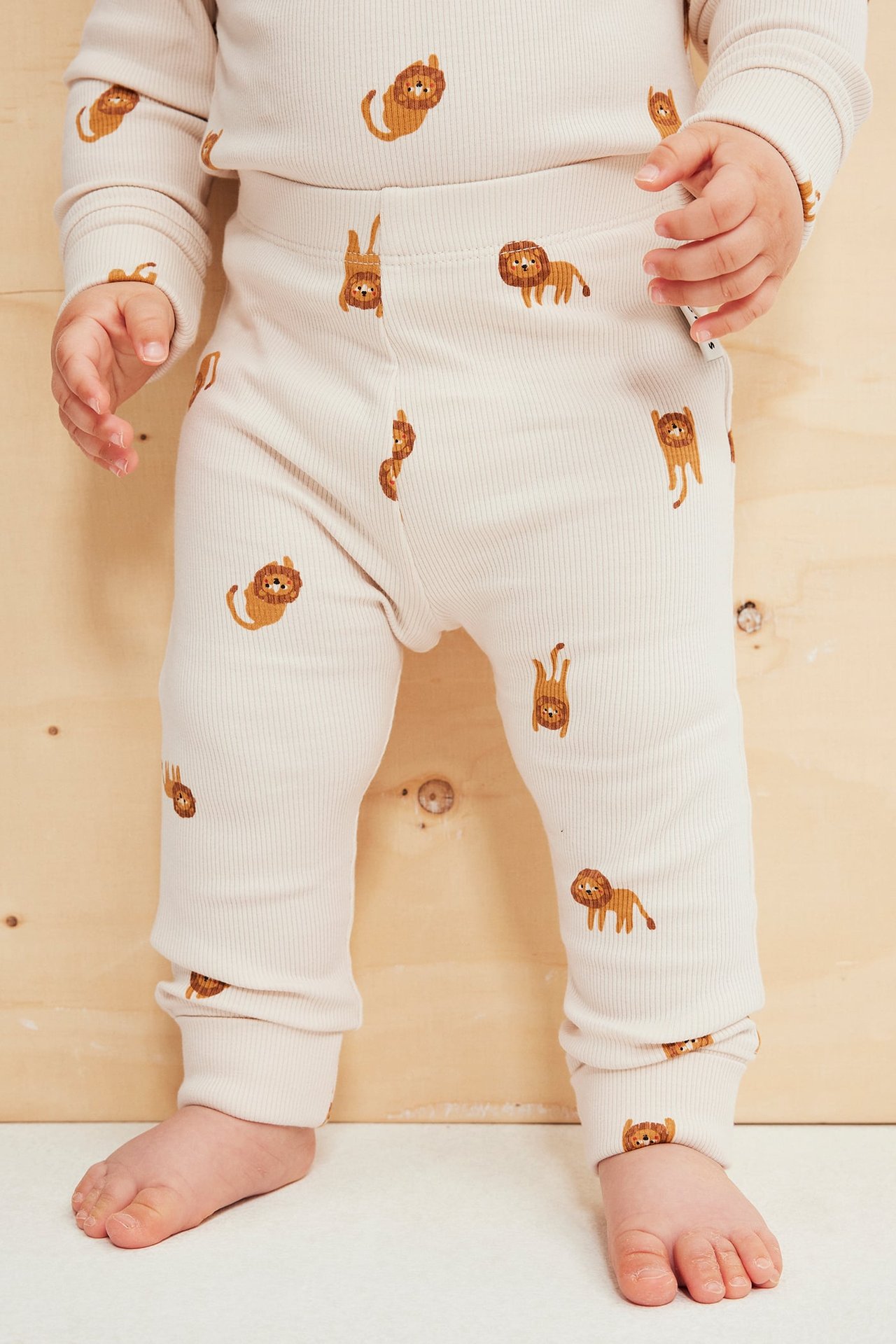 Forfra, baby i beige ribbestrikket langermet topp og matchende bukser med løvemønster.