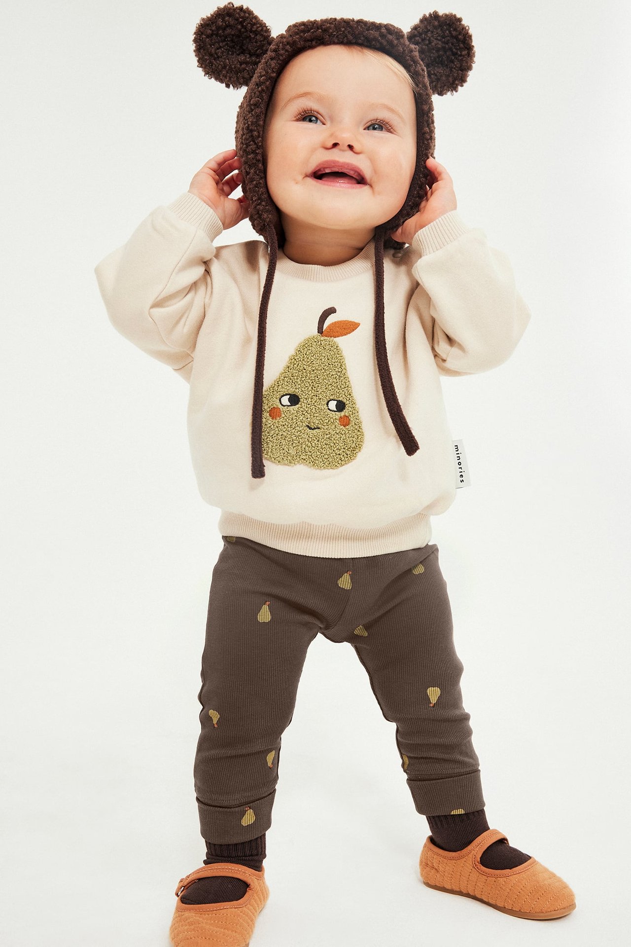 Framsida: baby i beige päronsweatshirt, bruna päronleggings, brun björnmössa och orange skor.