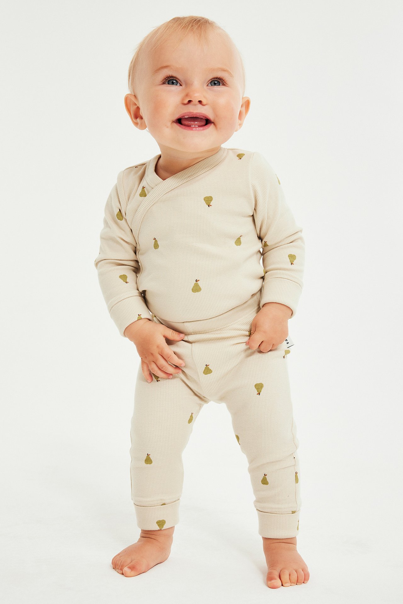 Frontbilde av baby i beige ribbet langermet omslagstopp og matchende leggings med grønt pæretrykk.