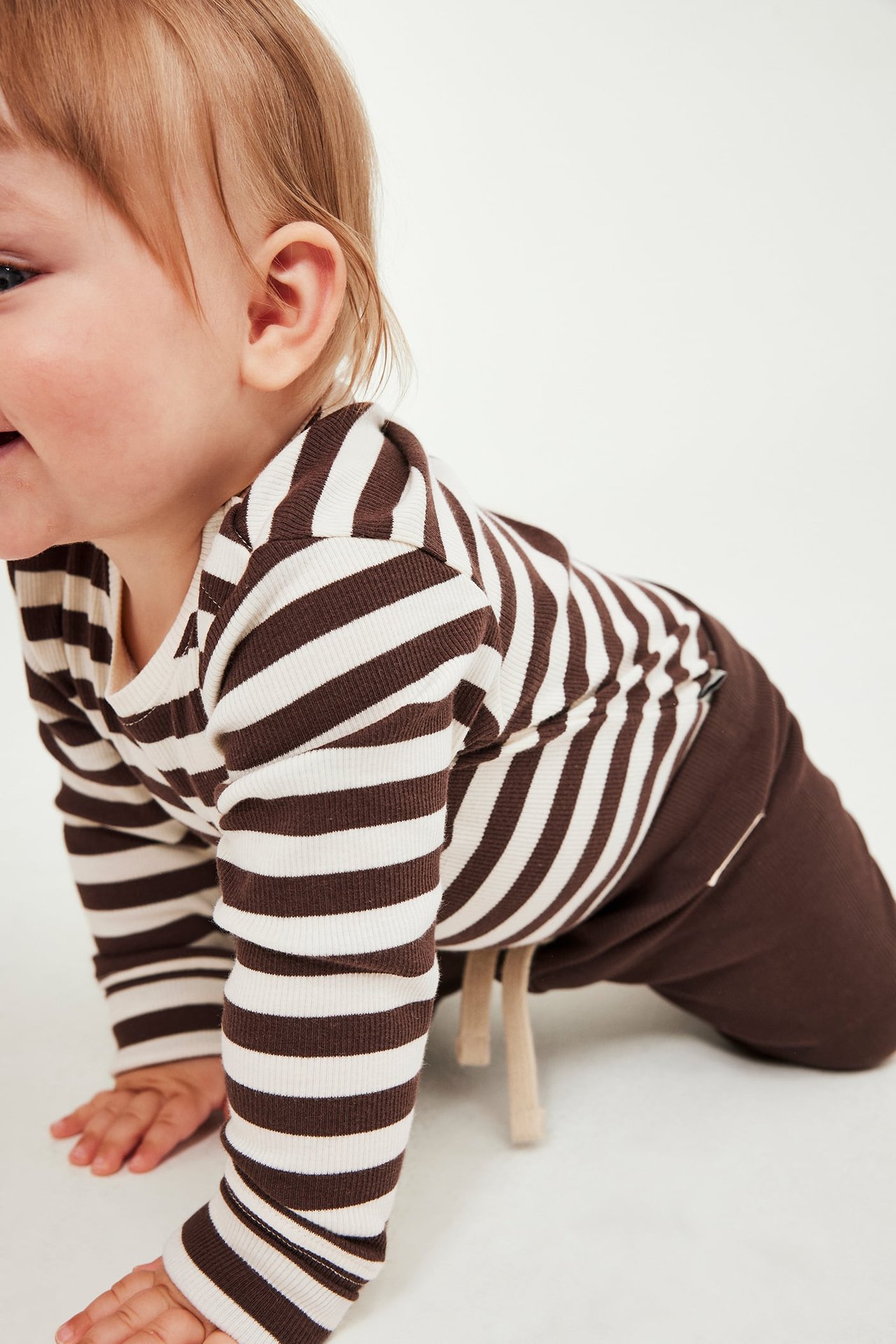 Sideutsikt av baby i brun og kremhvit stripet langermet topp og brune bukser.