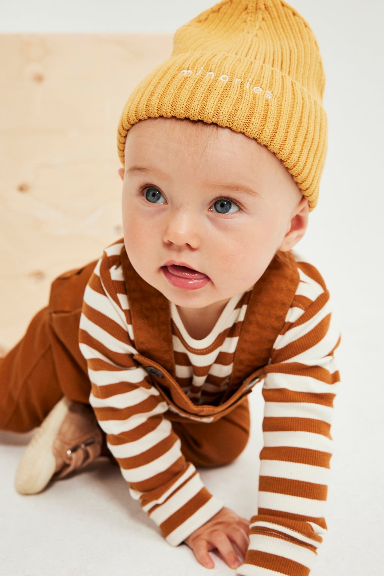 Krypende baby iført gul ribbestrikket lue, hvitstripet genser og brun selebukse.