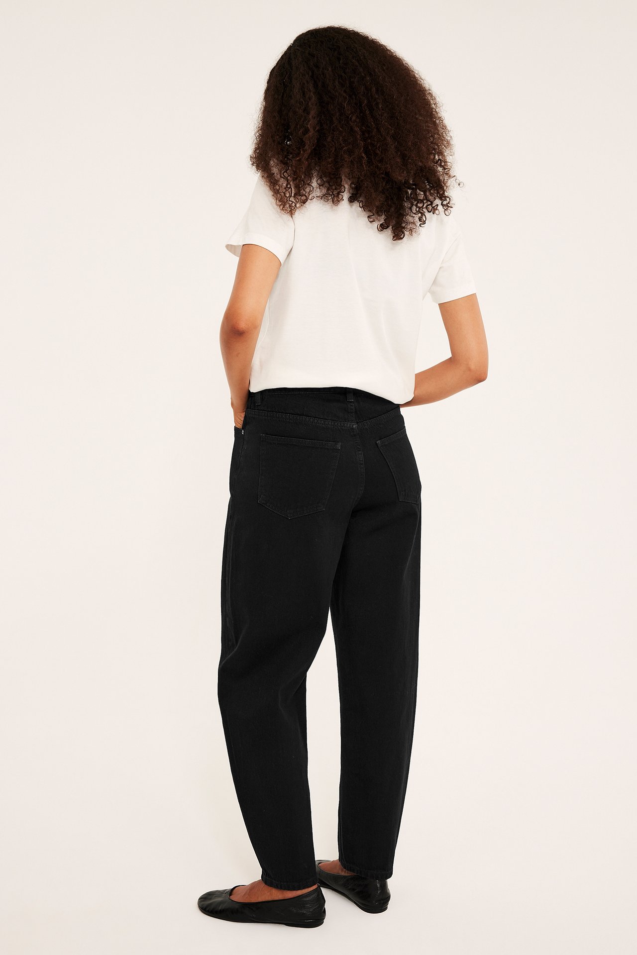 Widok od tyłu kobiety w czarnych jeansach relaxed fit, białym t-shircie i czarnych baletkach.
