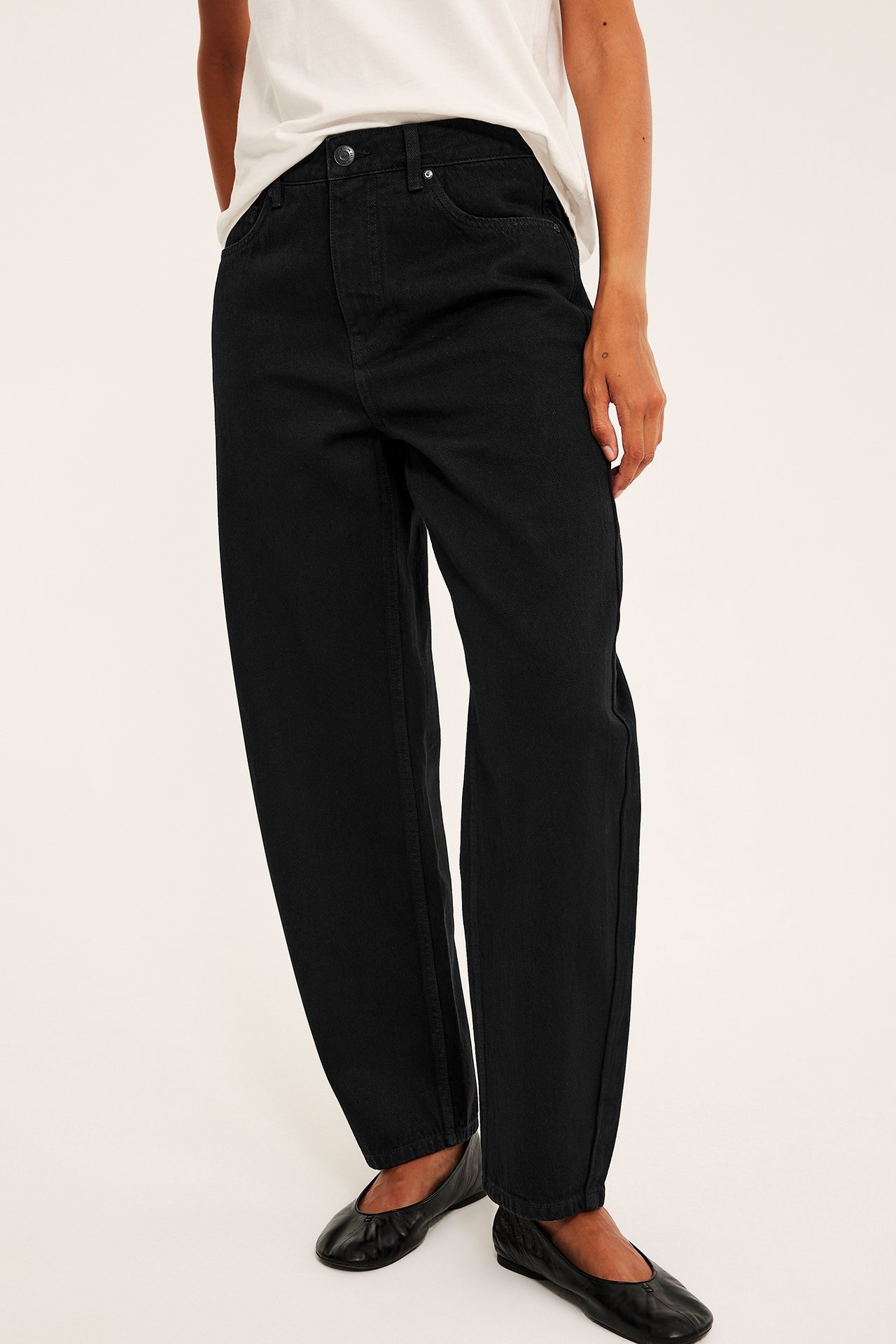 Forfra, kvinne iført svarte løse jeans med høy midje, hvit t-skjorte og svarte ballerinasko.
