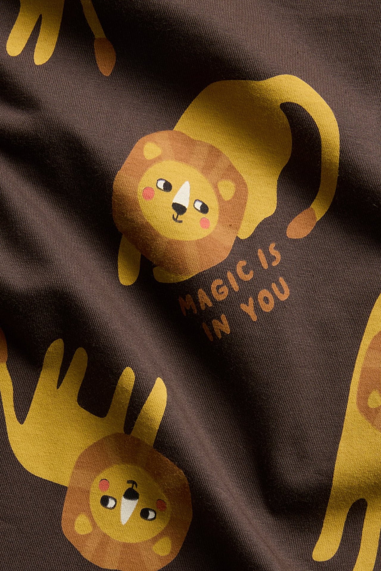 Zbliżenie na ciemnobrązowy materiał z powtarzającym się wzorem lwa z kreskówki i napisem 'MAGIC IS IN YOU'.