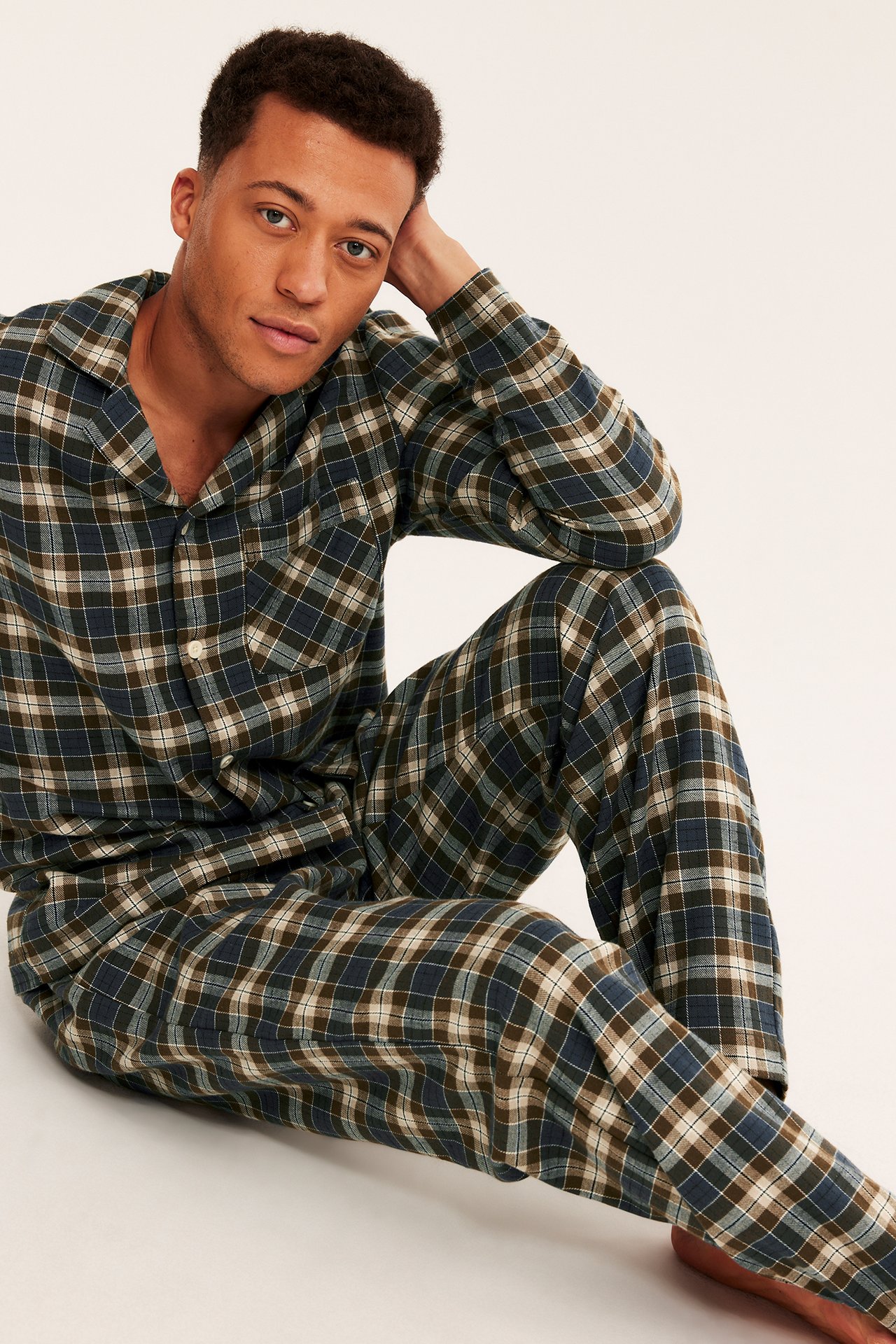 Framsida av sittande vuxen man i mörkblå, grön, brun och vitrutig pyjamas.