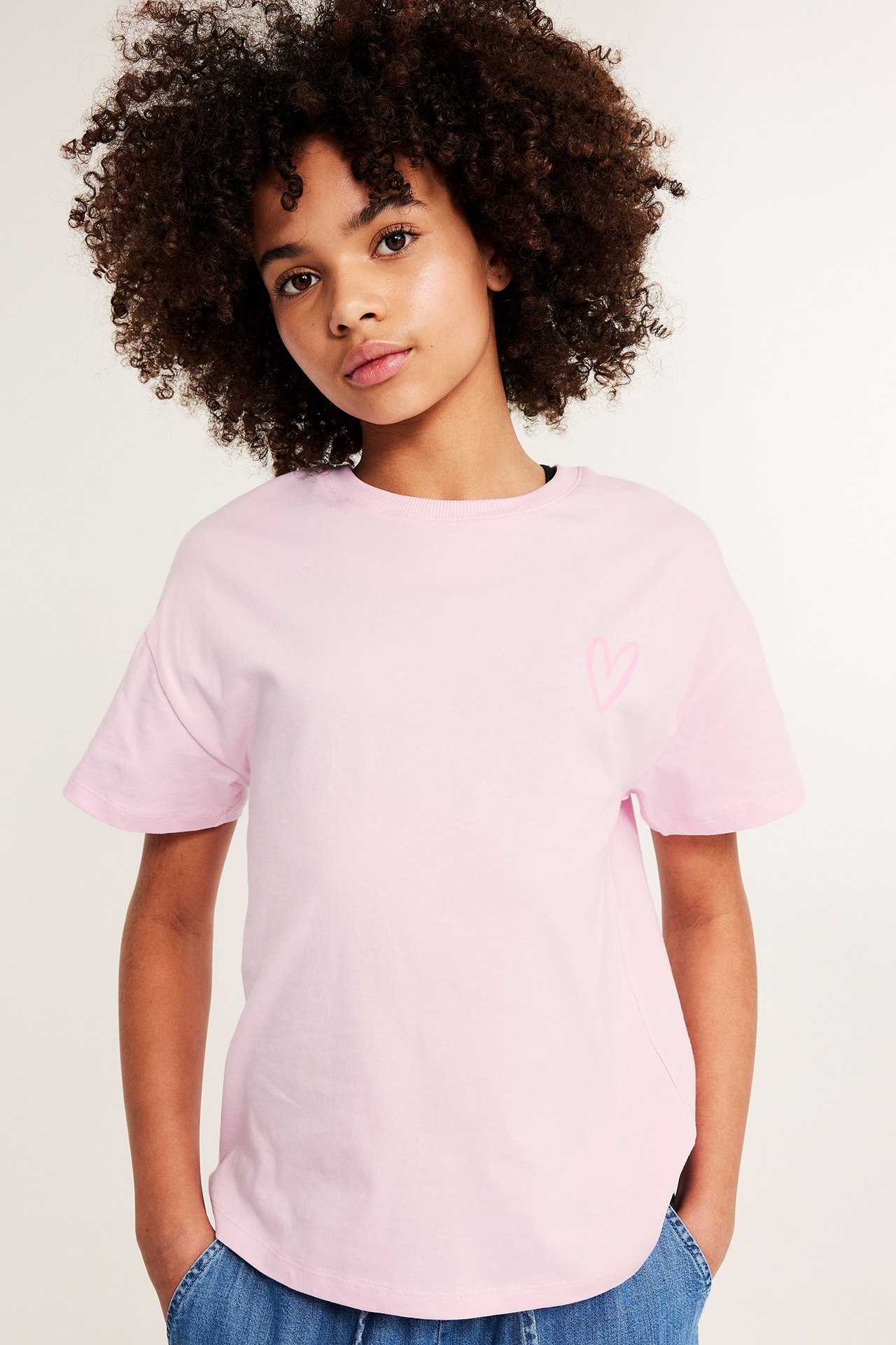 Edestä: Lapsi yllään vaaleanpunainen puuvilla-t-paita sydänkuviolla, sinisten farkkujen kanssa.