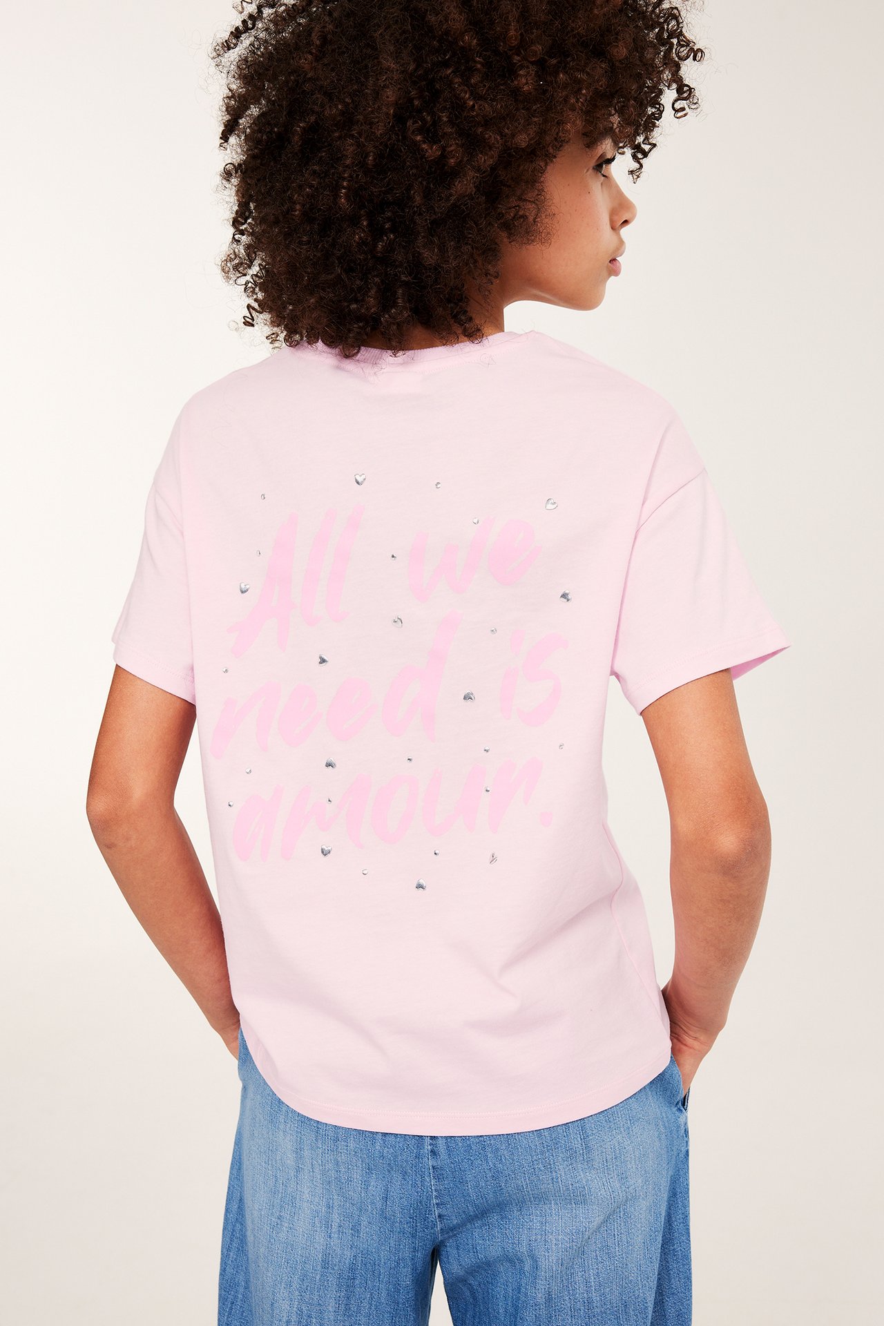 Bakfra, et barn i en rosa T-skjorte med teksttrykk, strass og blå dongeribukse.
