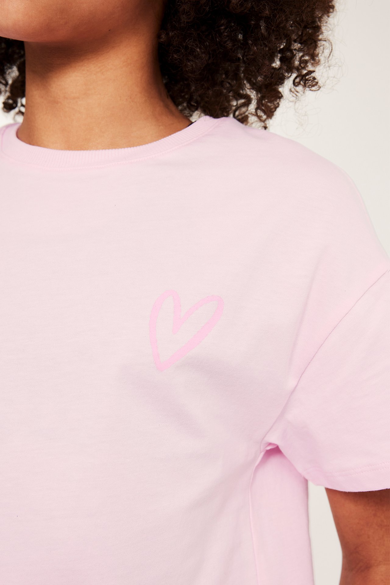 Widok z przodu: różowy bawełniany T-shirt z jasnoróżowym nadrukiem serca, noszony przez dziecko.