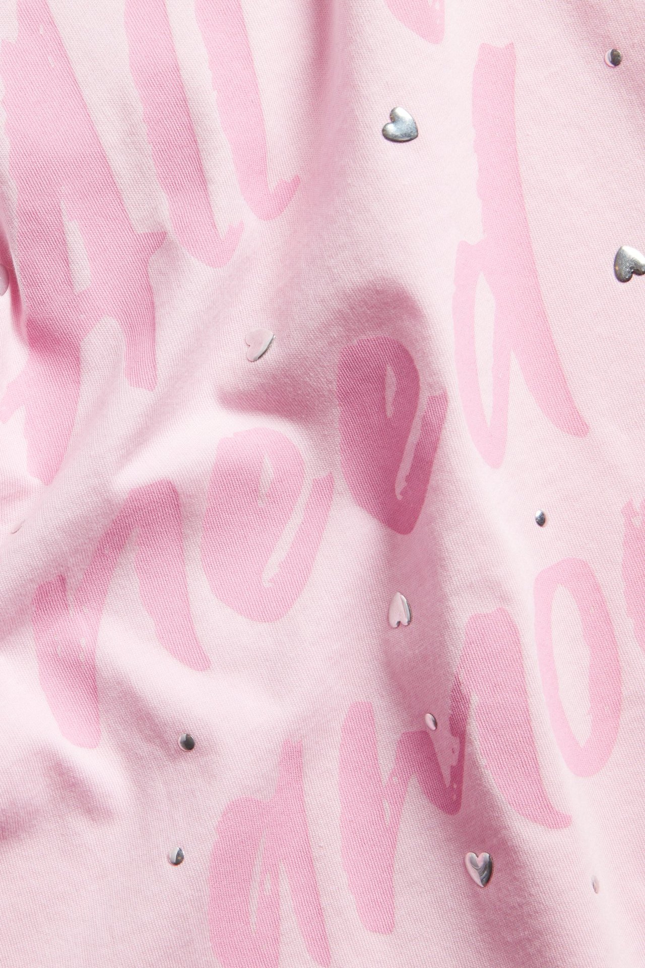 Närbild av en rosa T-shirt i bomull med ljusrosa texttryck och silverfärgade strasshjärtan.