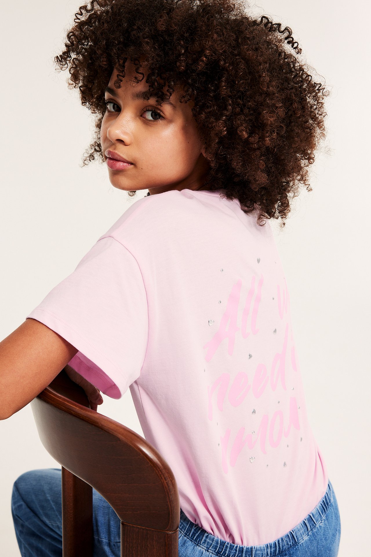 Takakuva lapsesta vaaleanpunaisessa puuvilla-T-paidassa strassipainatuksella, sinisten farkkujen kanssa.