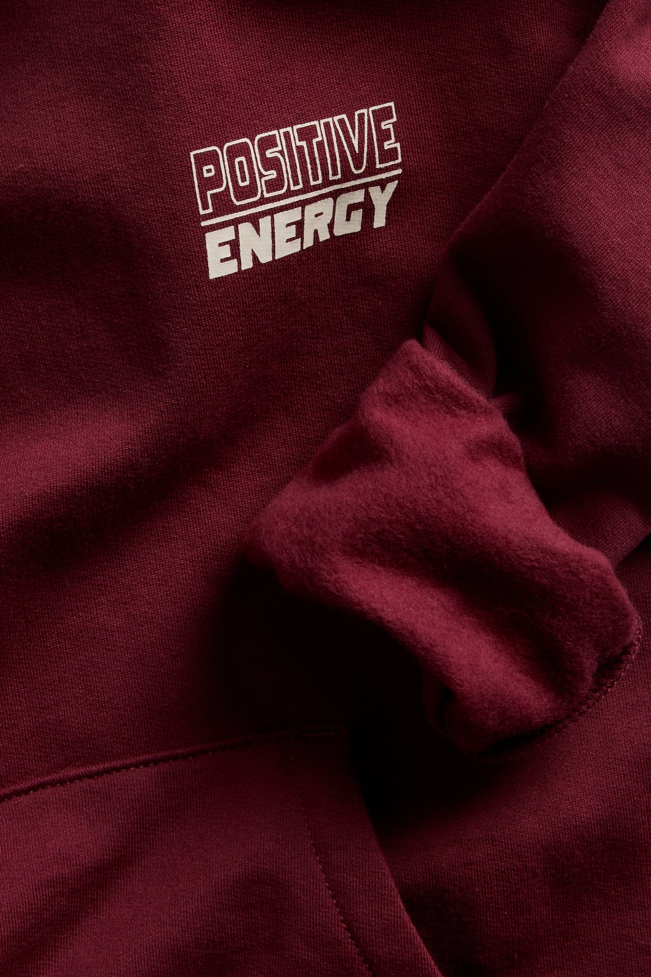 Zbliżenie na bordową bluzę z kapturem z białym napisem „POSITIVE ENERGY” na piersi.